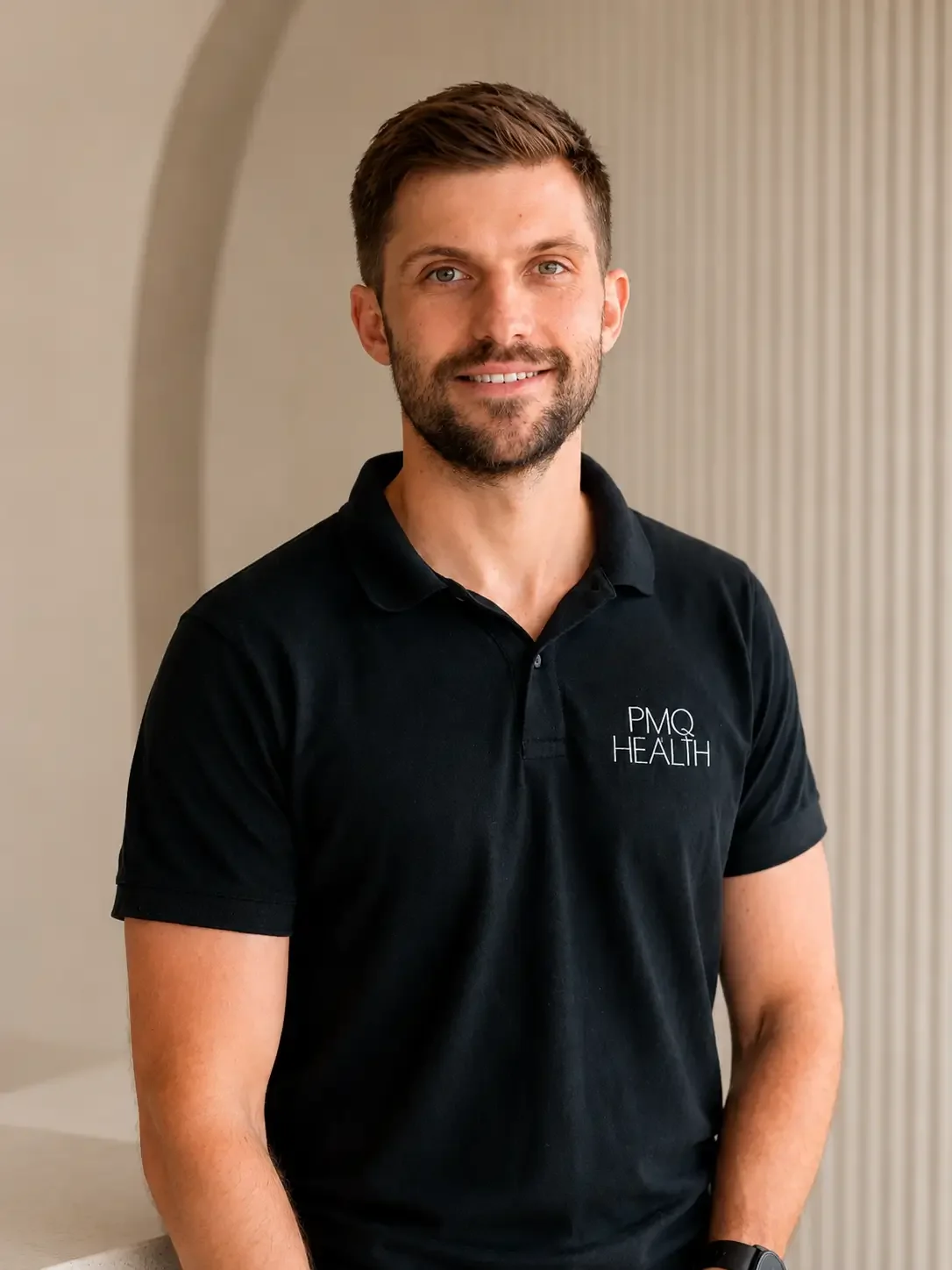 A man with short brown hair and a beard, wearing a black polo shirt with 'PMQ HEALTH' embroidered on it, standing indoors with a light-colored wall behind him.