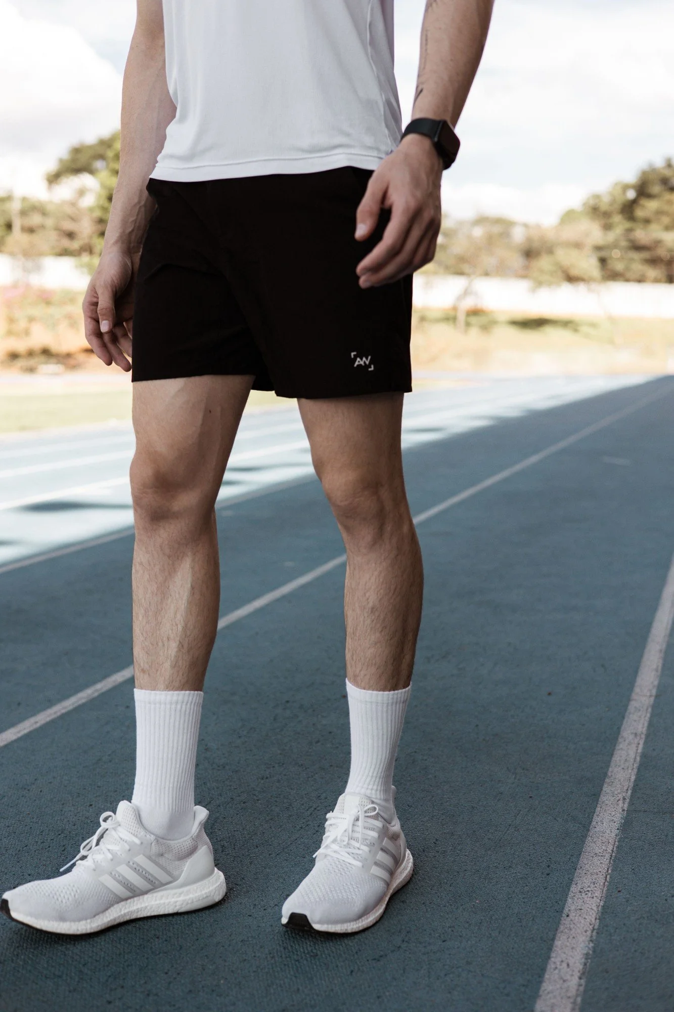 A person standing on a blue running track wearing white sneakers, white socks, black shorts, a white t-shirt, a black wristwatch, and white socks. The background shows trees and a cloudy sky.