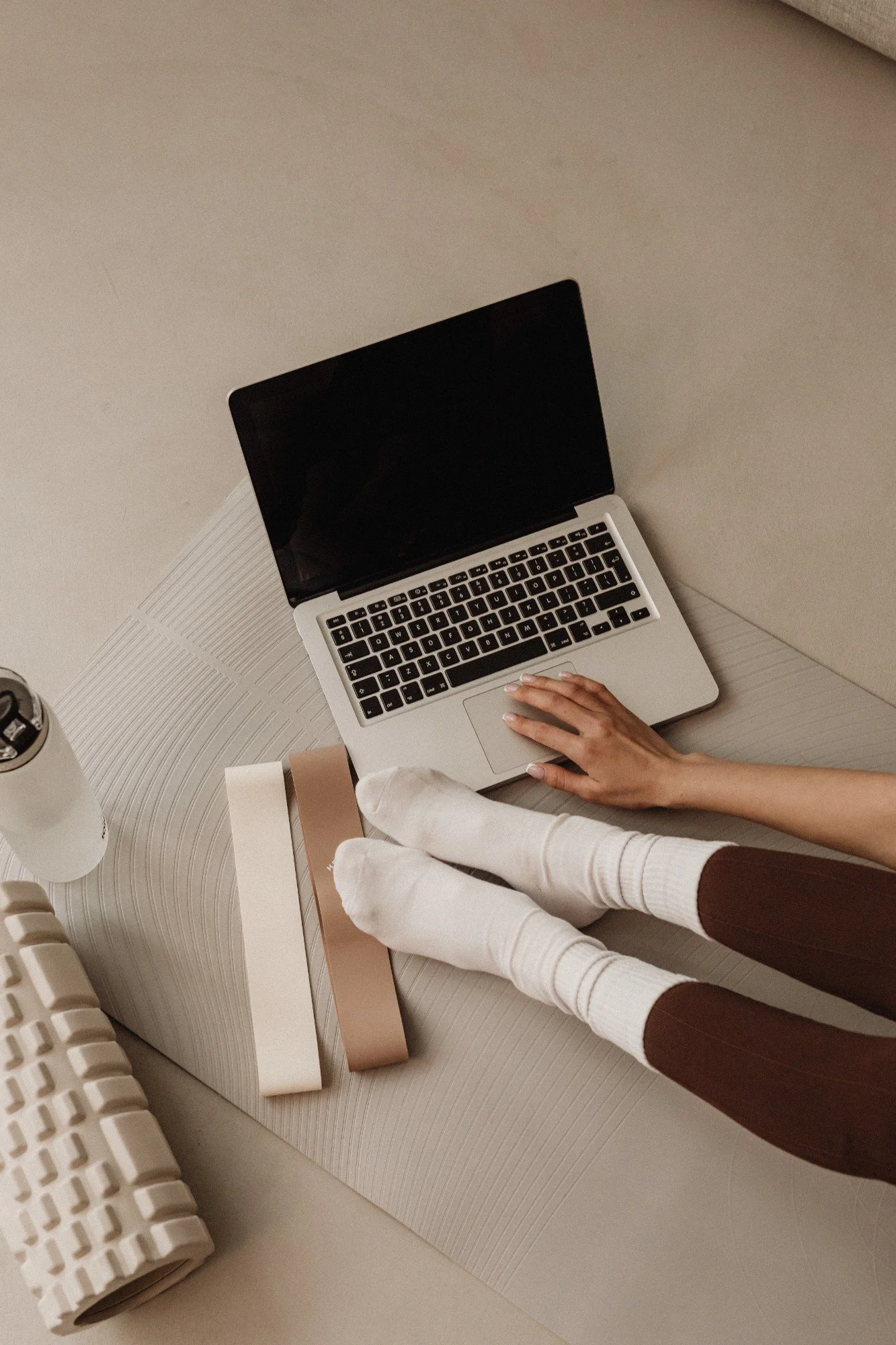A person with dressed in white and brown sleeve long pants sitting on a beige textured surface, using a silver laptop with a black screen. There are various rectangular and cylindrical objects nearby, including a white keyboard and a white container with a lid.