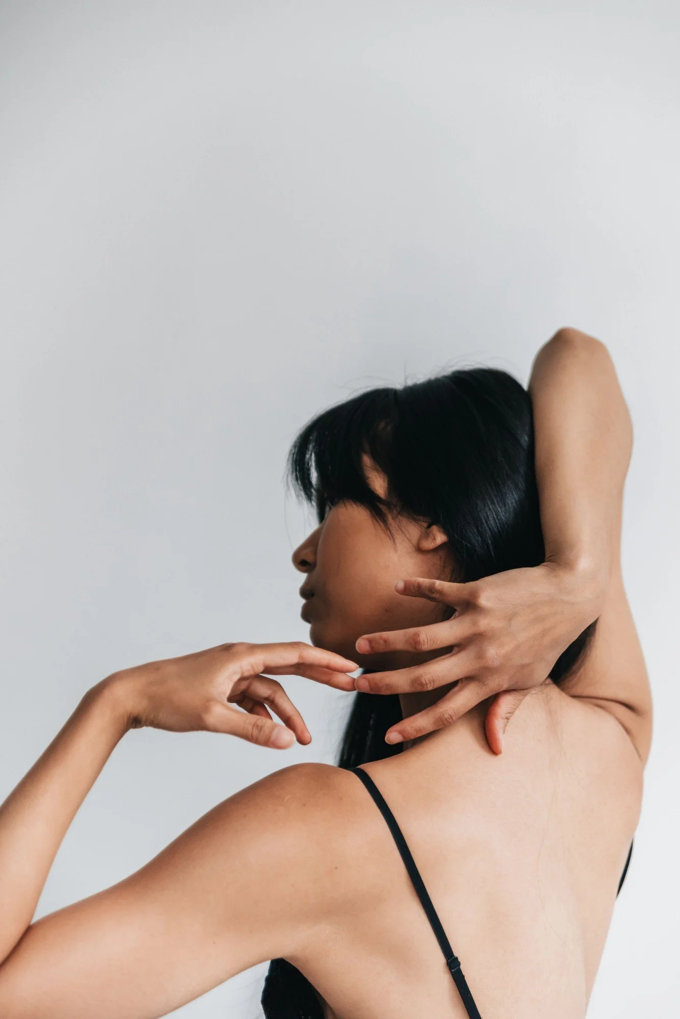 A woman with black hair and wearing a black spaghetti strap top is posing with her eyes closed, her right hand is gently touching her face near her chin, and her left hand is behind her head, with her head turned slightly to the right. The background is plain and light-colored.