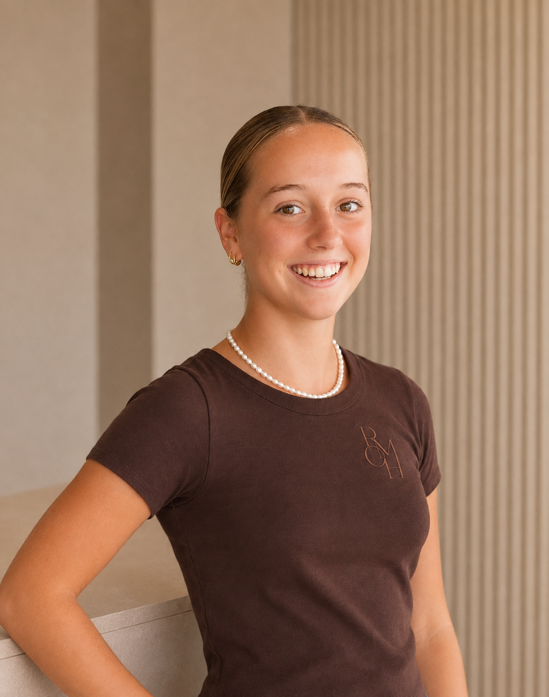 A young woman smiling, wearing a brown T-shirt, pearl necklace, and hoop earrings, standing indoors against a neutral background.
