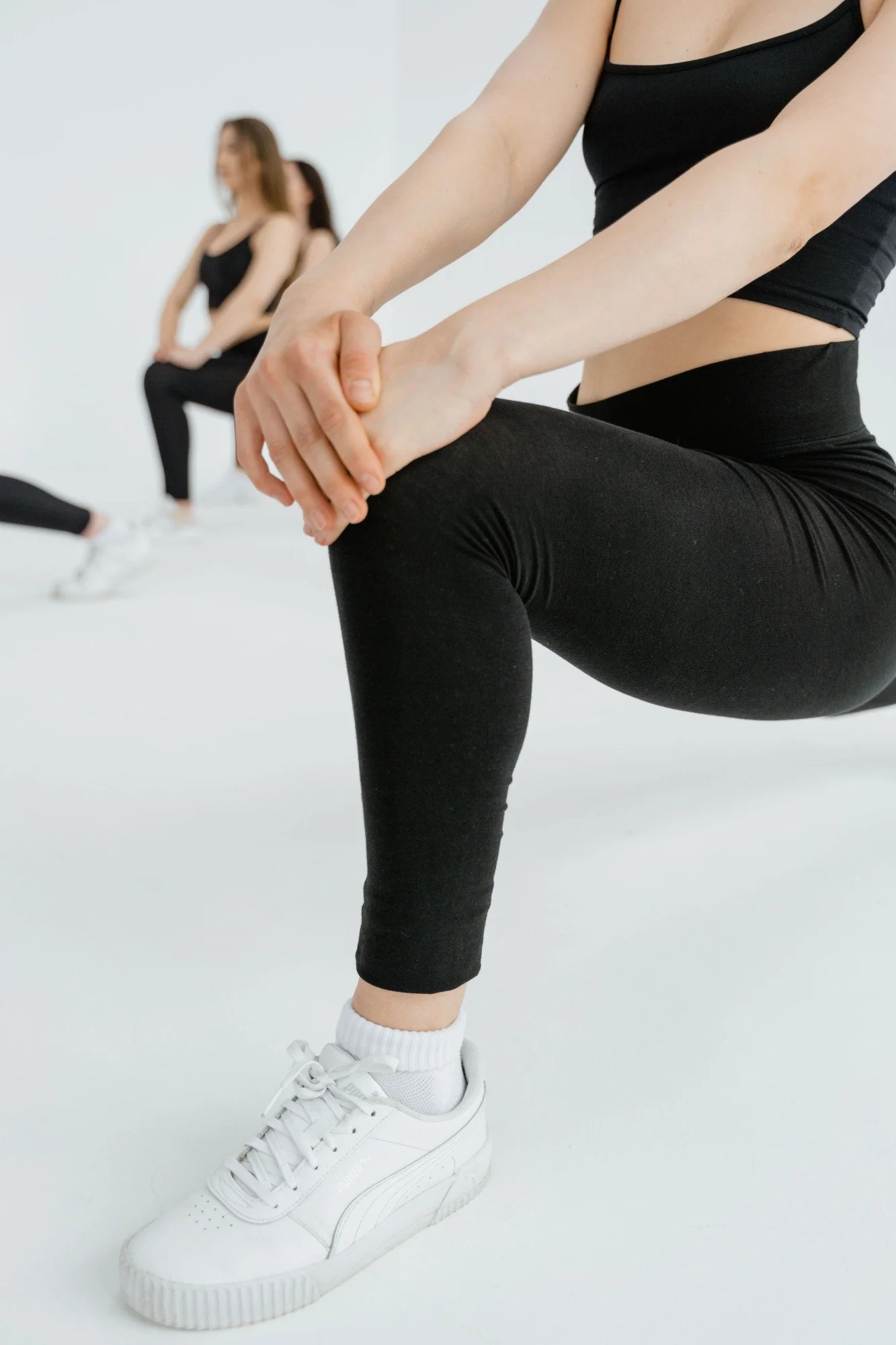 A person in black workout clothes doing a squat with hands clasped in front. In the background, others are also stretching or exercising. The setting has a white floor and wall.