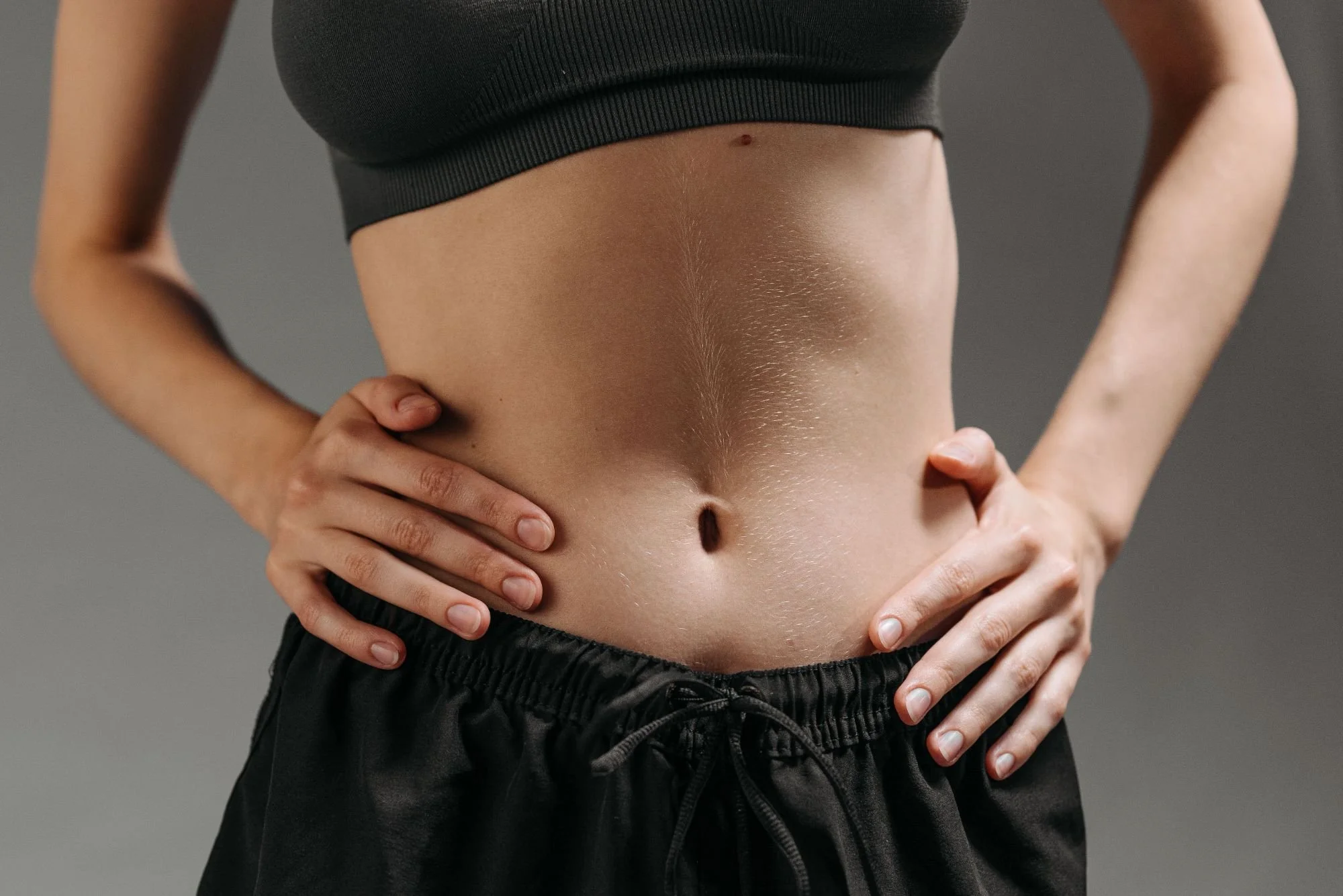 Close-up of a person's bare midsection with hands positioned on hips, wearing a black crop top and black pants, against a gray background.