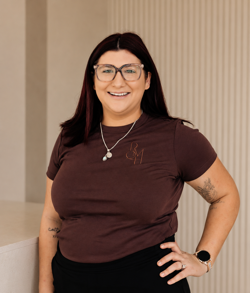 A woman with dark red hair, glasses, and a brown T-shirt smiling and standing with her hand on her hip indoors.