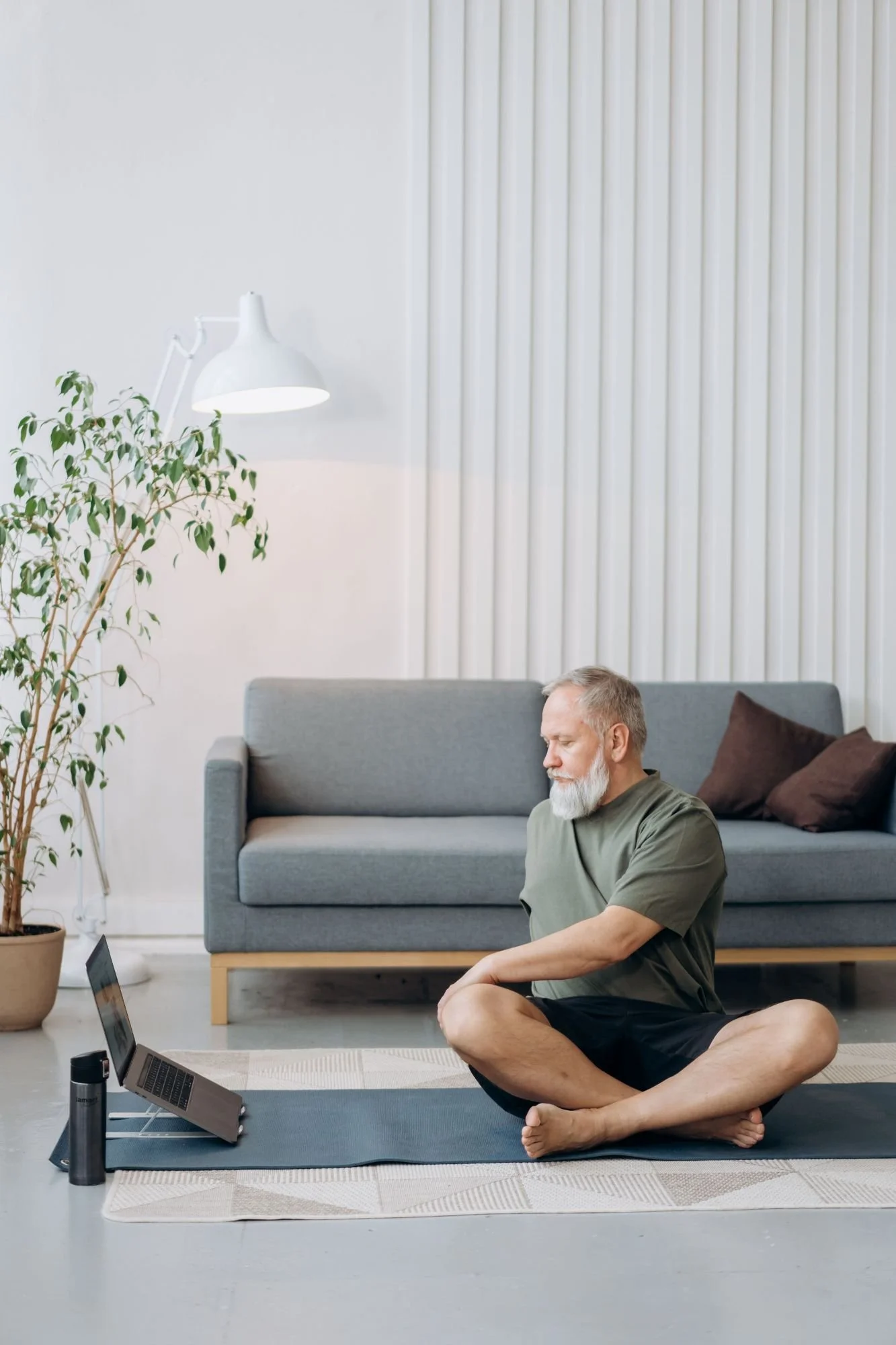 A man with white hair and beard, sitting on a yoga mat in a living room, meditating with closed eyes in front of a laptop. The room features a gray sofa, a large potted plant, a white wall with vertical blinds, and a white wall lamp.