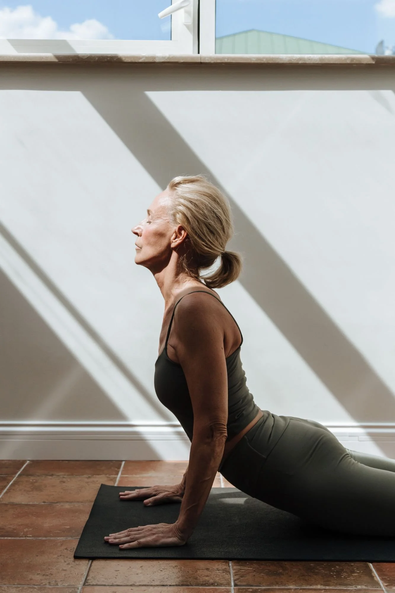 A woman performing downward dog yoga pose indoors, with sunlight and shadows cast on the wall behind her.