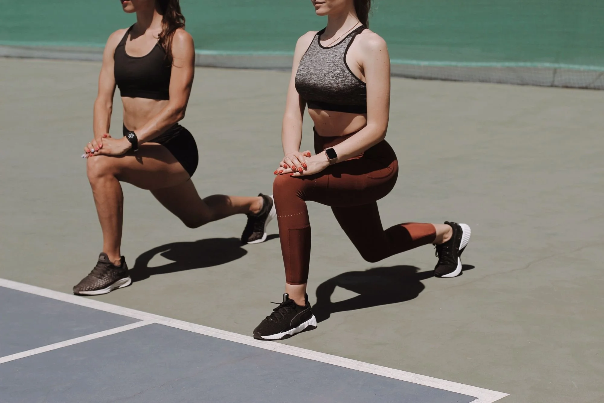 Two women practicing yoga outdoors on a sunny day near the water, each in a kneeling lunge position with hands together in a prayer gesture.
