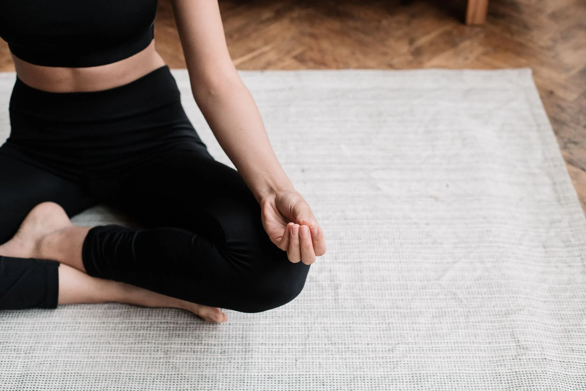 A person practicing yoga or meditation on a mat indoors, sitting cross-legged with their right hand resting on their knee, making a mudra gesture with their thumb and index finger.