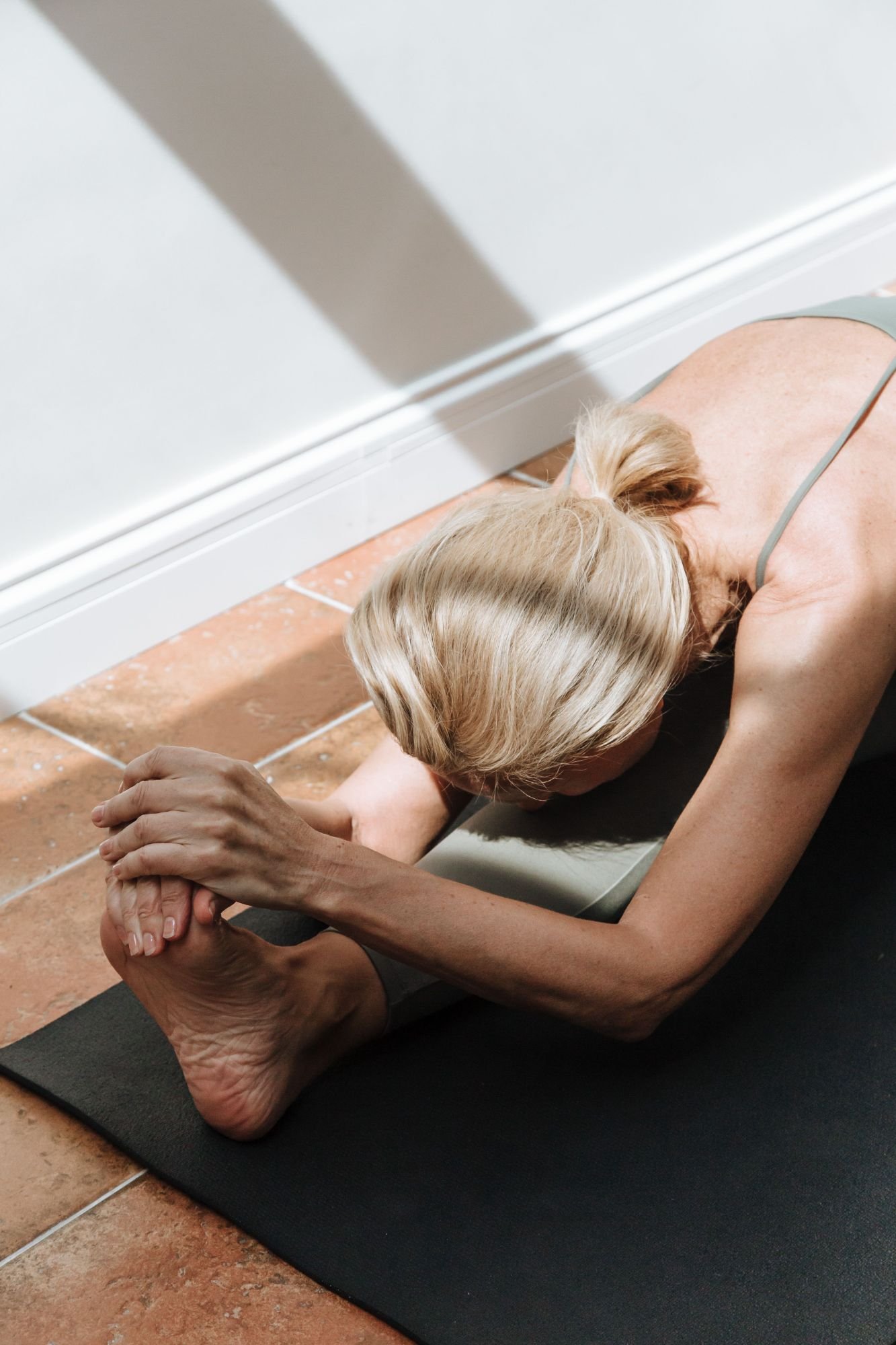 An elderly woman with blonde hair lying on a yoga mat in a yoga pose, holding her feet with her hands, practicing yoga indoors.