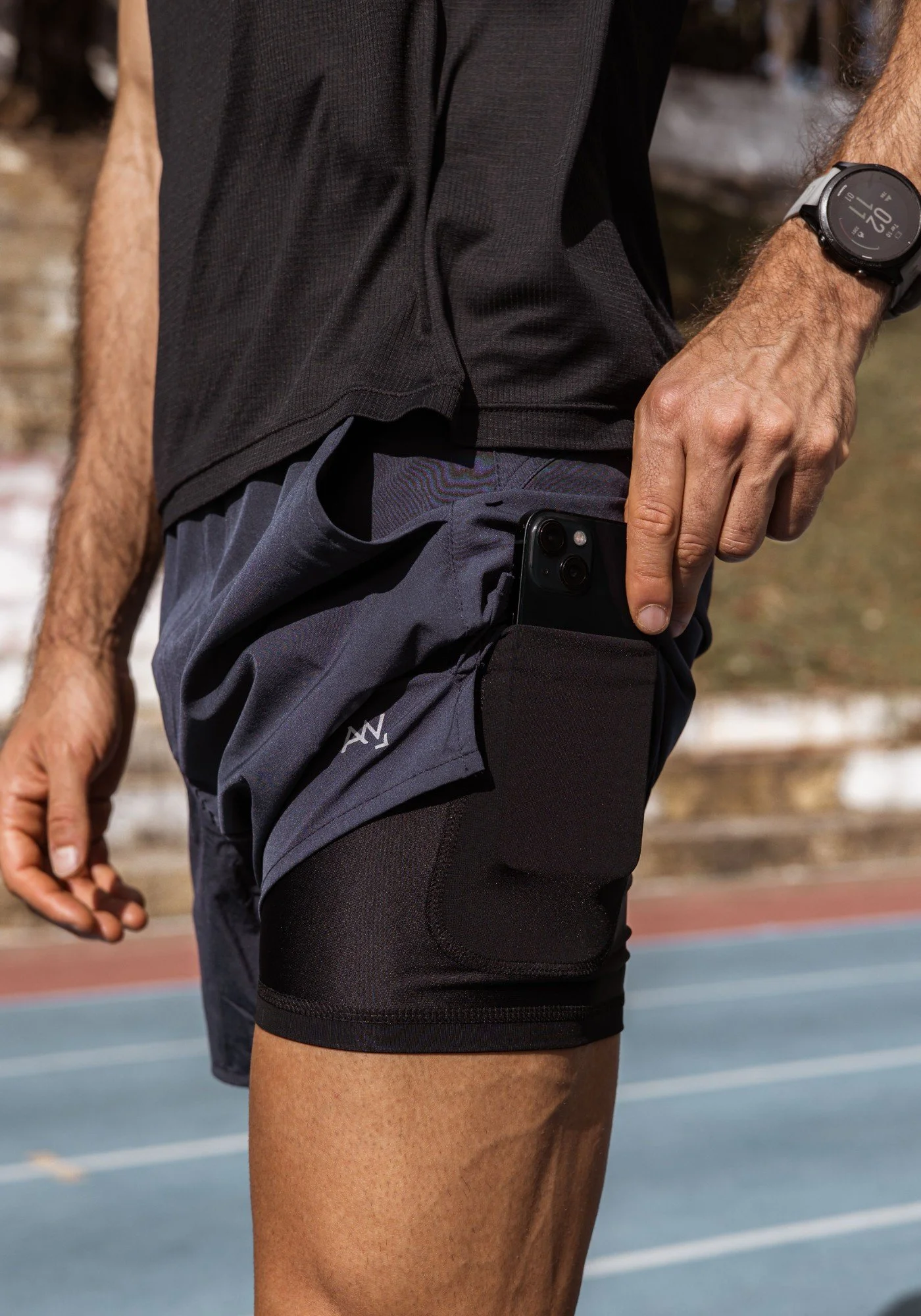 A person wearing a black sports shirt, black shorts with a pocket, a wristwatch, and holding a smartphone in a hand. The person is standing on a running track outdoors.