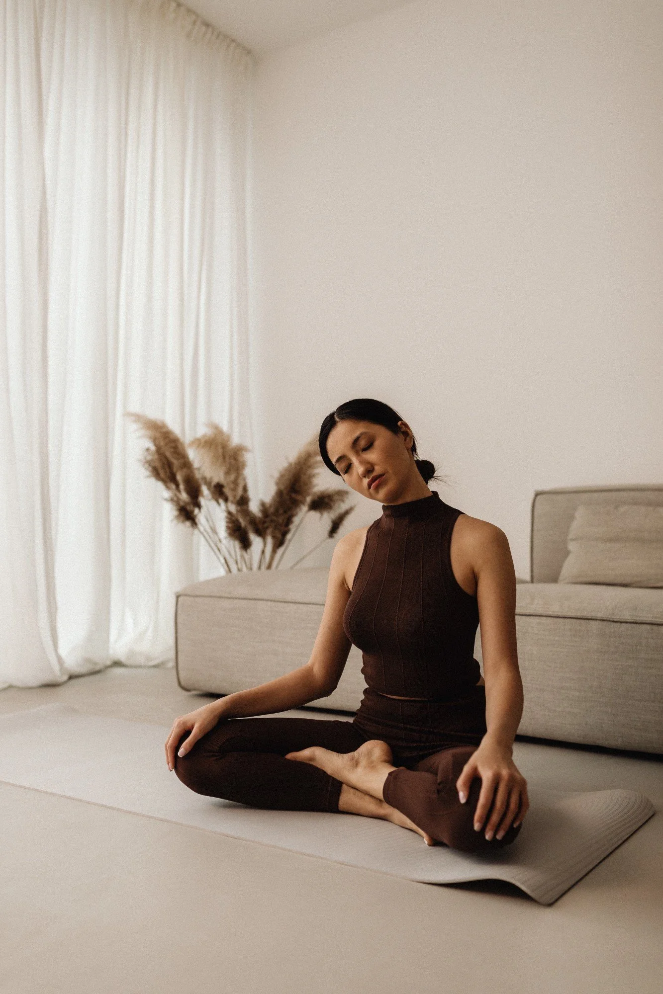A woman practicing yoga in a minimalist living room, sitting cross-legged on a yoga mat, with eyes closed and a calm expression, beige sofa with pampas grass in the background.