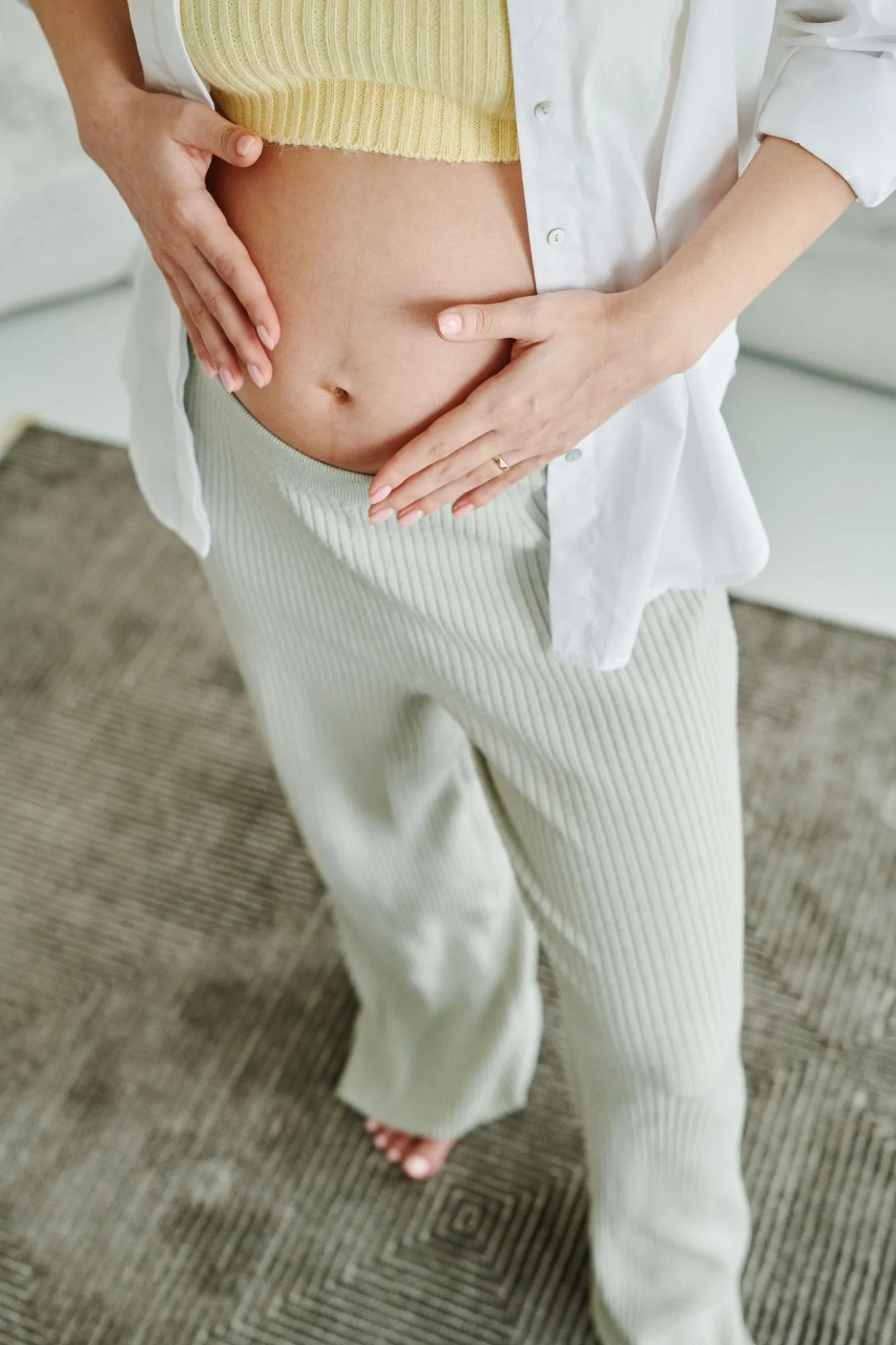 A person wearing a yellow top, white unbuttoned shirt, and beige striped pants standing on a patterned carpet. Their hands are placed on their bare stomach, with one hand on each side.