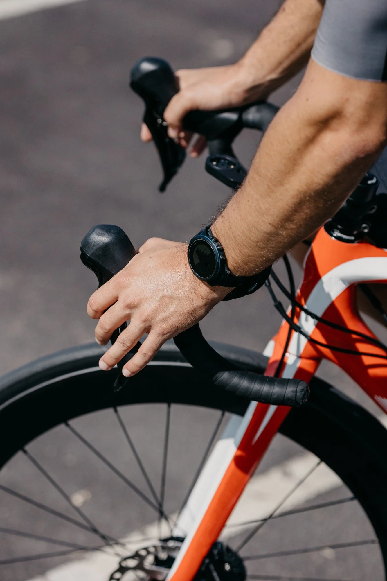 Close-up of a person riding a bicycle, showing their hands on the handlebars, a wristwatch, and part of the orange and white bicycle frame.