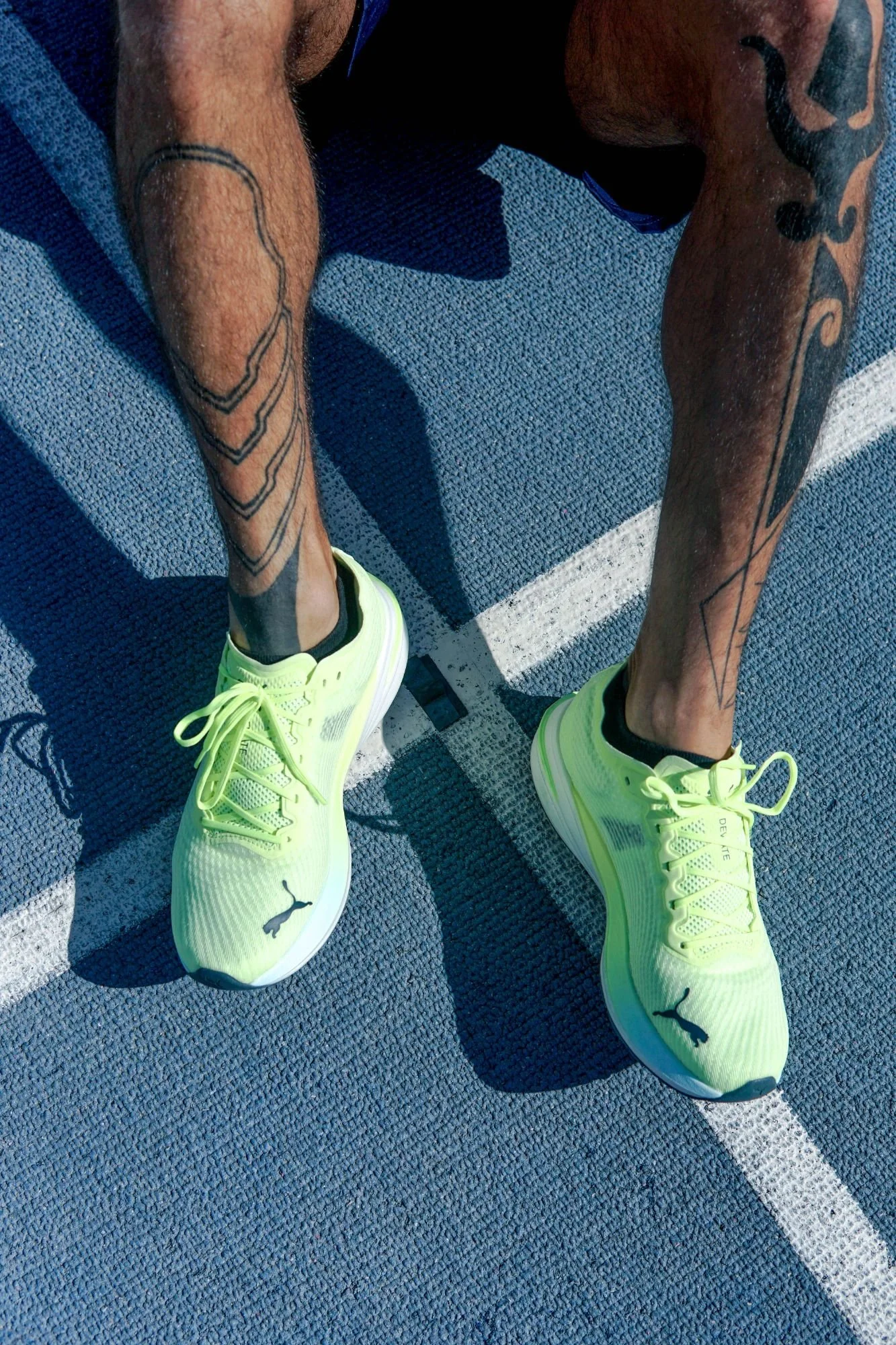 Close-up of a person wearing bright yellow Puma running shoes, black ankle socks, and shorts, standing on a blue running track. The person's arms and legs have visible tattoos.