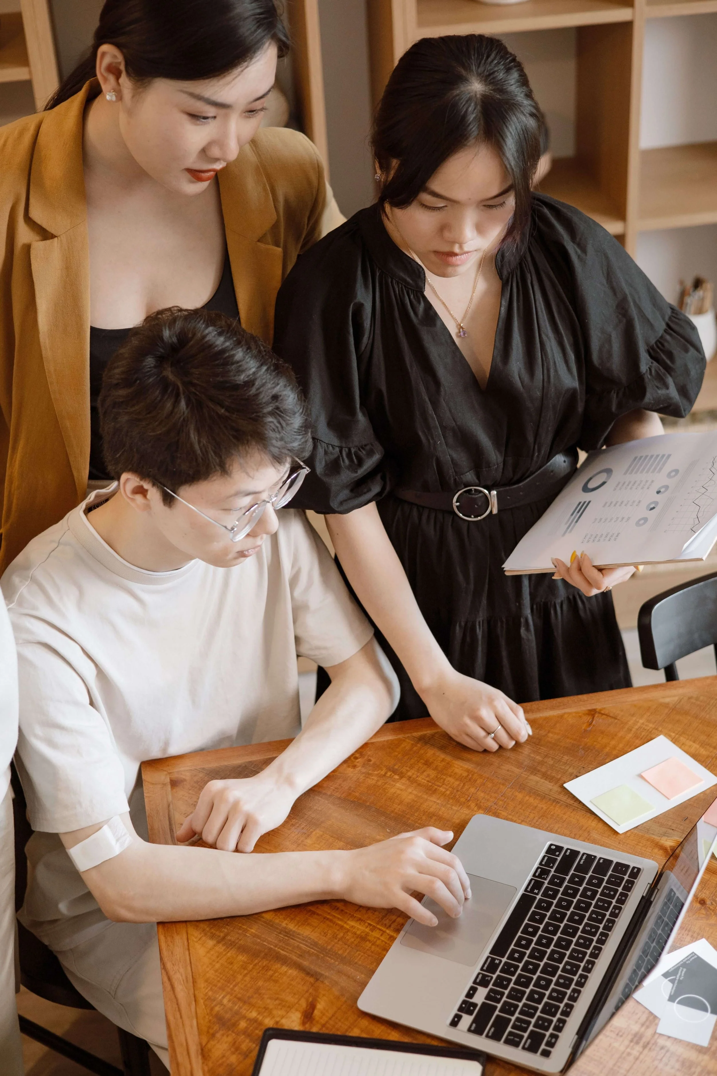Three women collaborating around a laptop and documents on a wooden table in an office or meeting room.