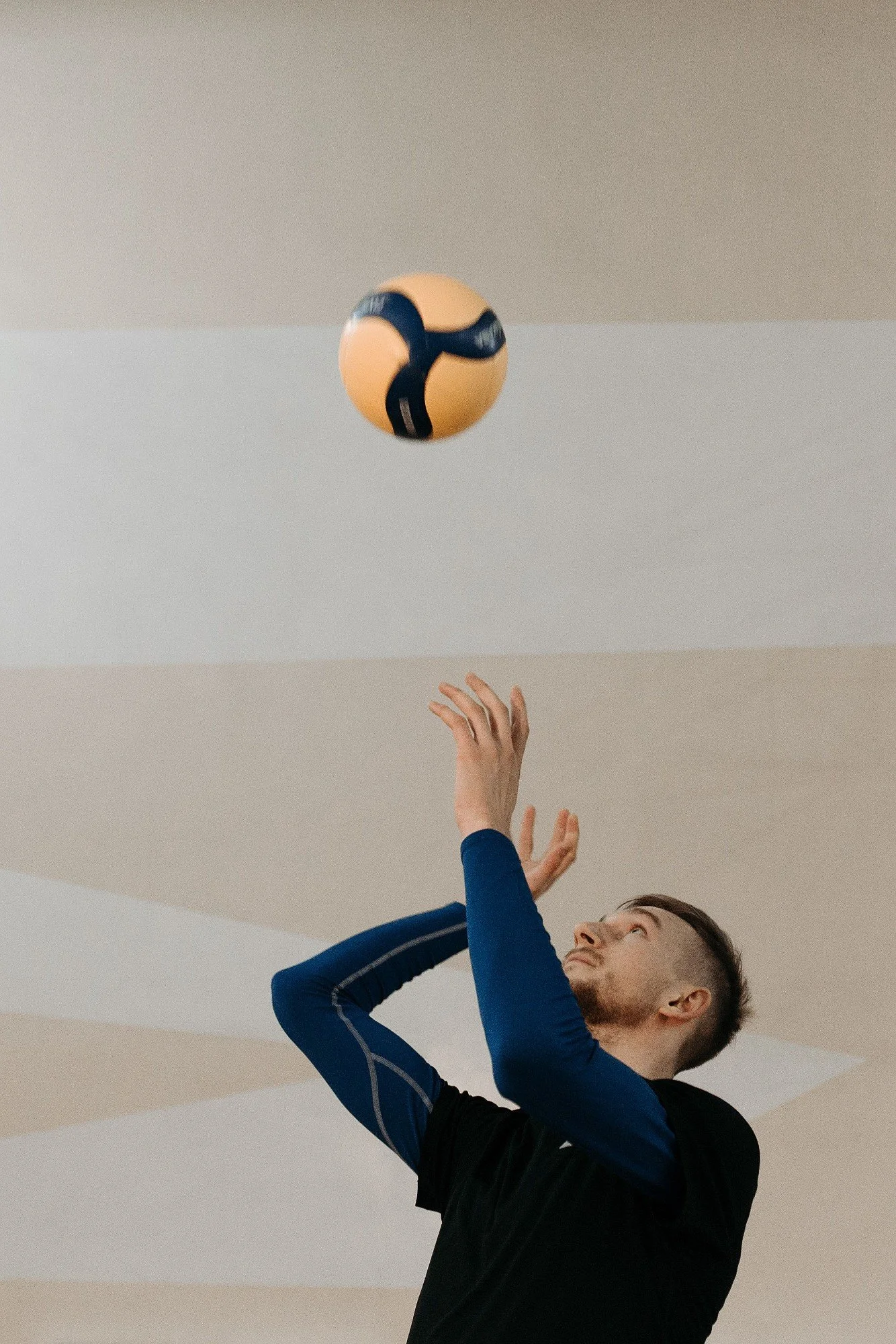 Young man in athletic clothing reaching up to hit a volleyball indoors.
