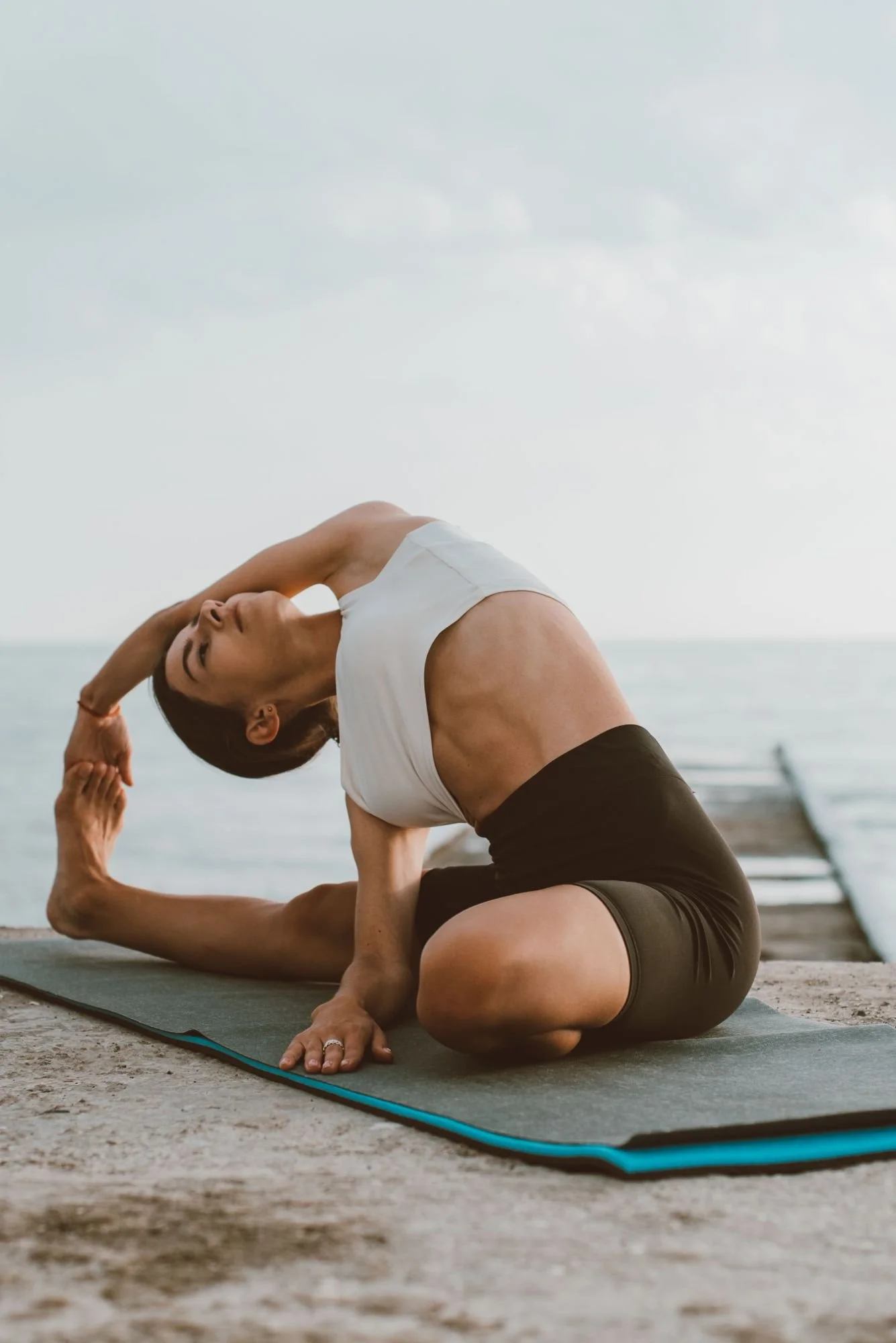 A woman practicing yoga outdoors on a mat by the water, performing a side bend stretch with her left arm over her head and her right arm supporting her on the ground.