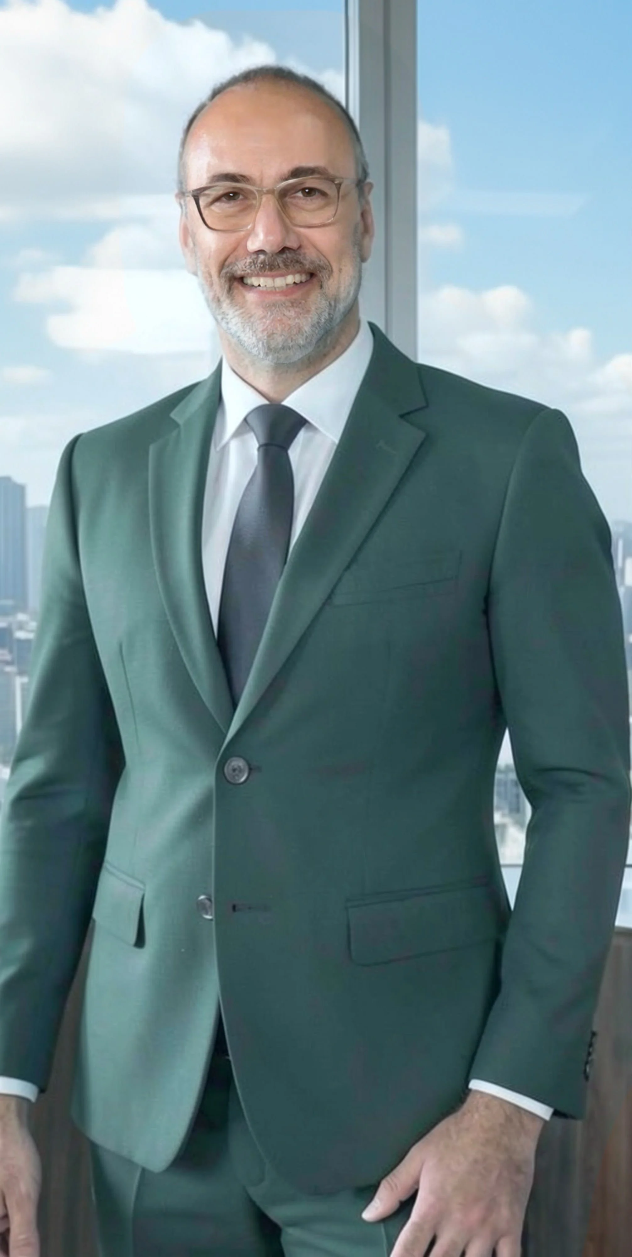 A middle-aged man with short hair, glasses, and a beard, wearing a dark green suit, white shirt, and black tie, standing indoors with a cityscape visible through the window behind him.