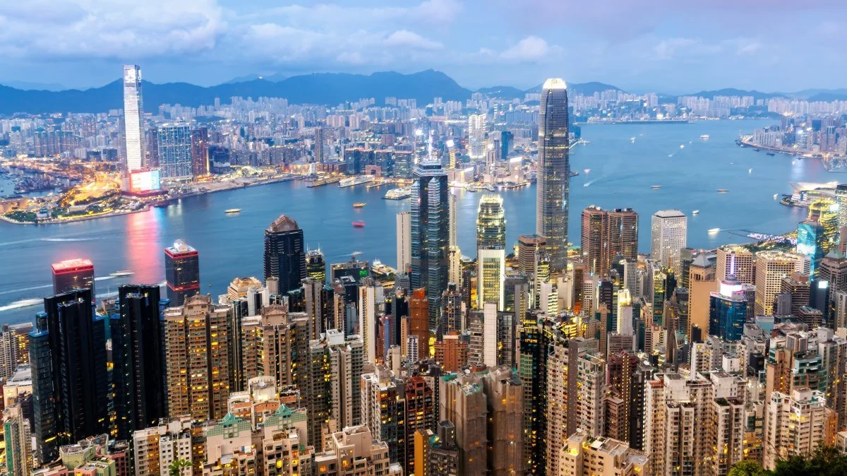 A panoramic view of Hong Kong's skyline at dusk with tall skyscrapers, Victoria Harbour, and mountain background.