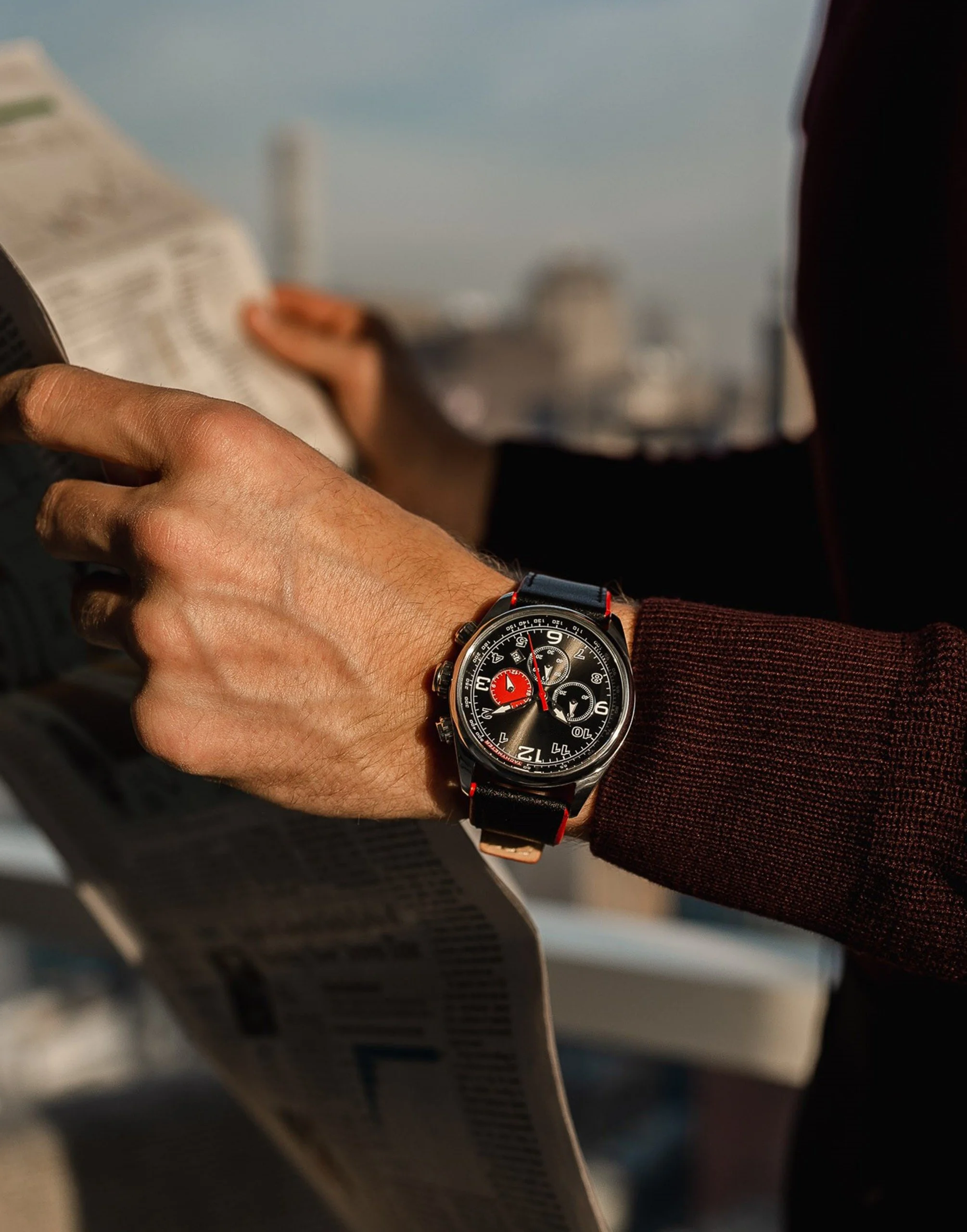 A person's hand is holding a newspaper, and they are wearing a black wristwatch with a large face, red accents, and multiple dials. The person is wearing a maroon sweater or jacket sleeve. In the background, there are blurred objects, possibly on a boat or outdoor platform, with a cloudy sky.