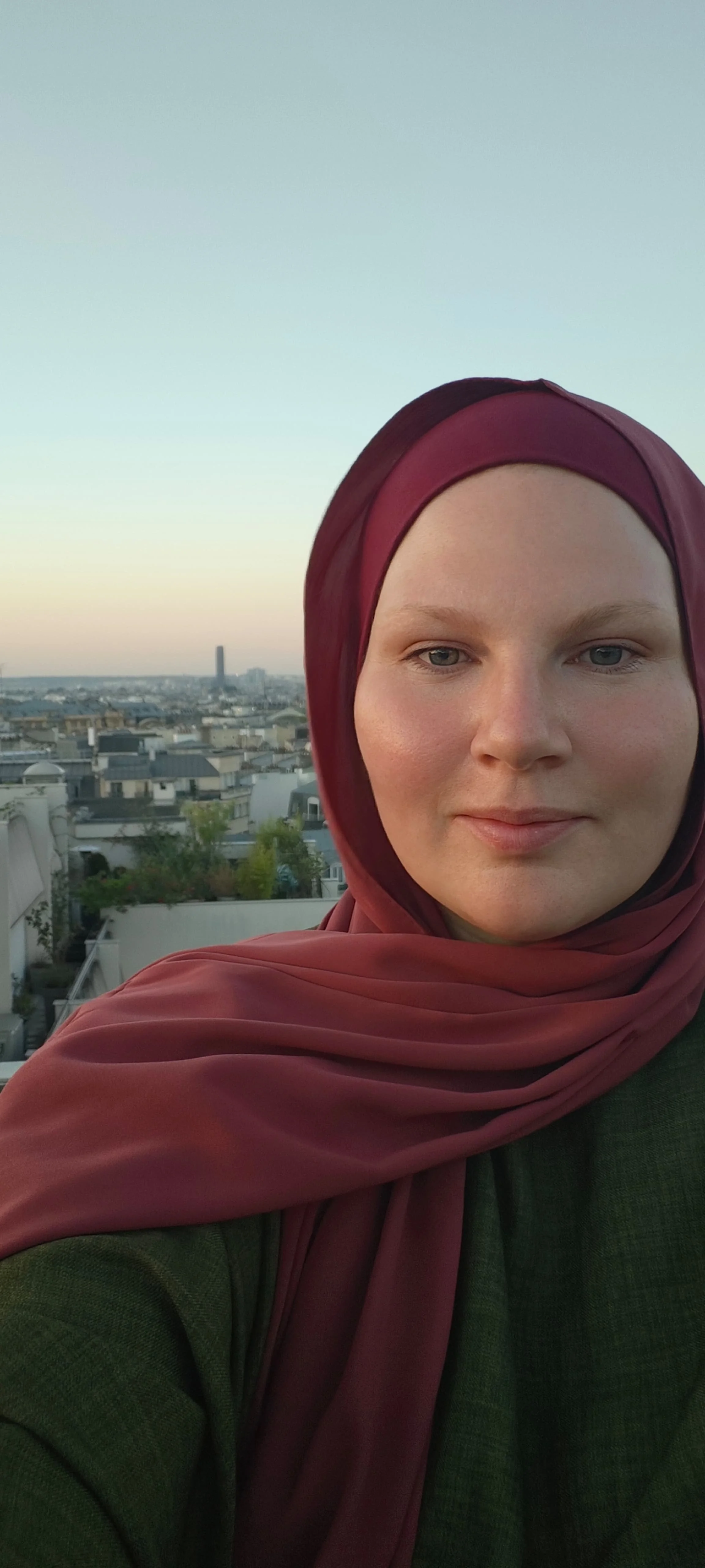 Une femme portant un hijab bordeaux, faisant un selfie avec une vue sur Paris au coucher du soleil.