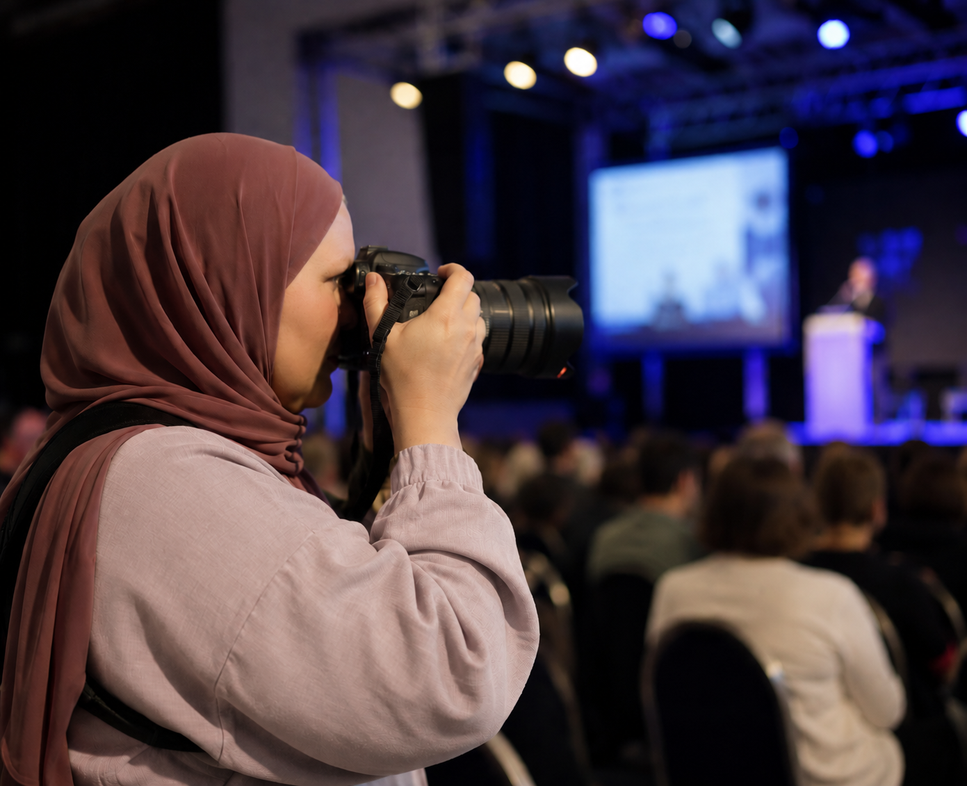 Femme portant un hijab marron prenant une photo lors d'une conférence avec un micro sur scène et un public en arrière-plan.