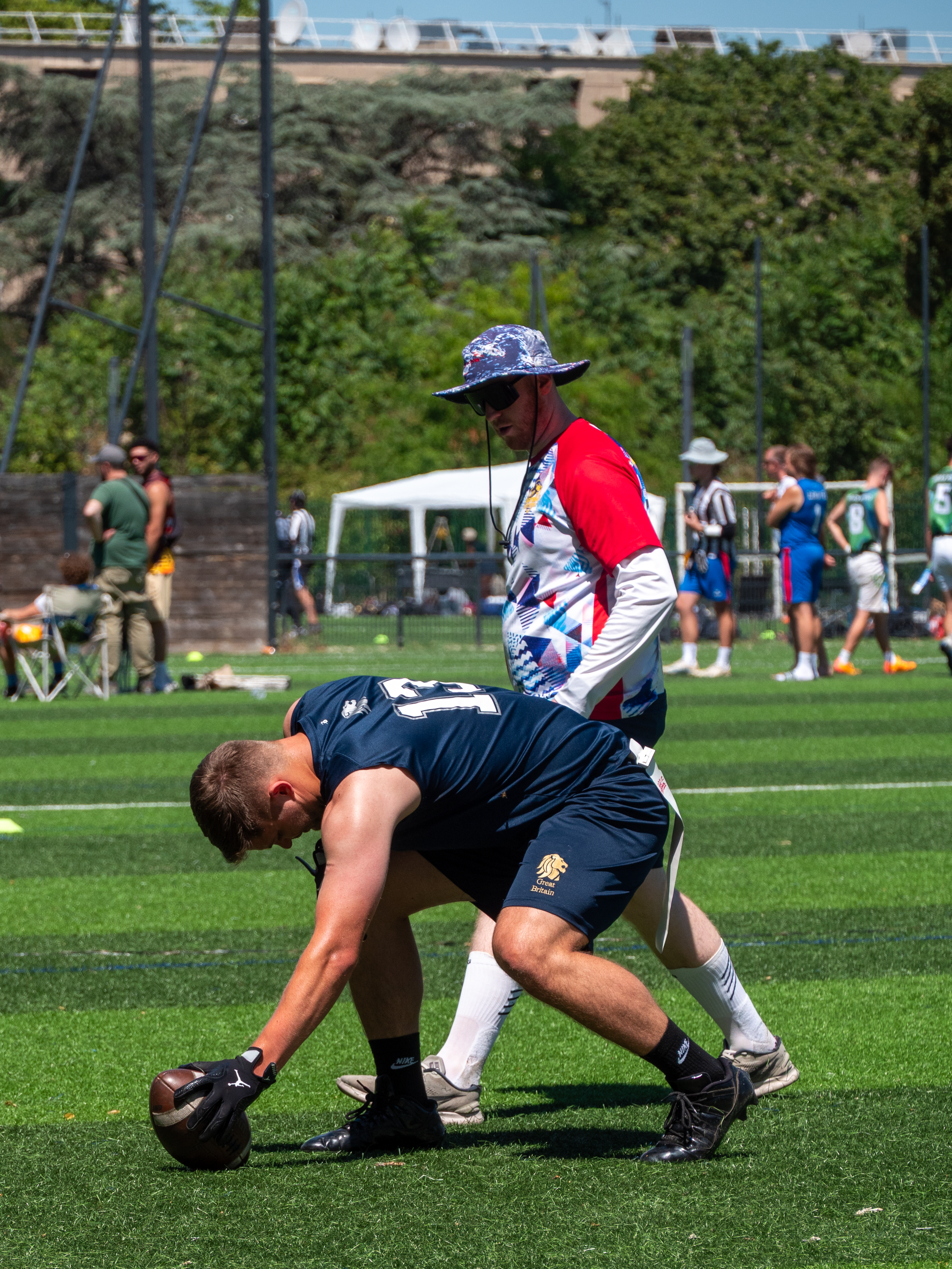 Deux joueurs de football américain s'affrontent lors d'un match, l'un d'eux est à genoux et attrape le ballon, tandis que l'autre, portant un chapeau à fleurs et des lunettes de soleil, se tient derrière lui sur un terrain herbeux avec des spectateurs en arrière-plan.