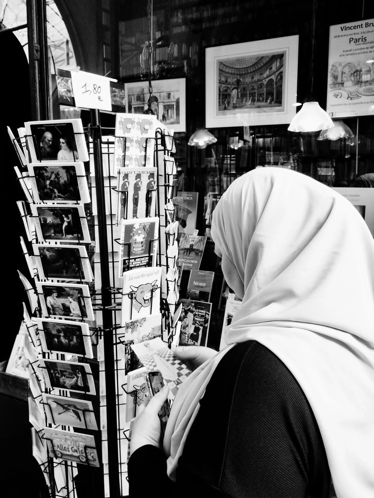 Une personne, en partie visible, portant un hijab blanc et un haut noir, regarde un panneau de cartes postales dans une librairie ou un magasin à Paris. Des affiches encadrées de peintures sont visibles derrière, ainsi qu'une affiche d'exposition.