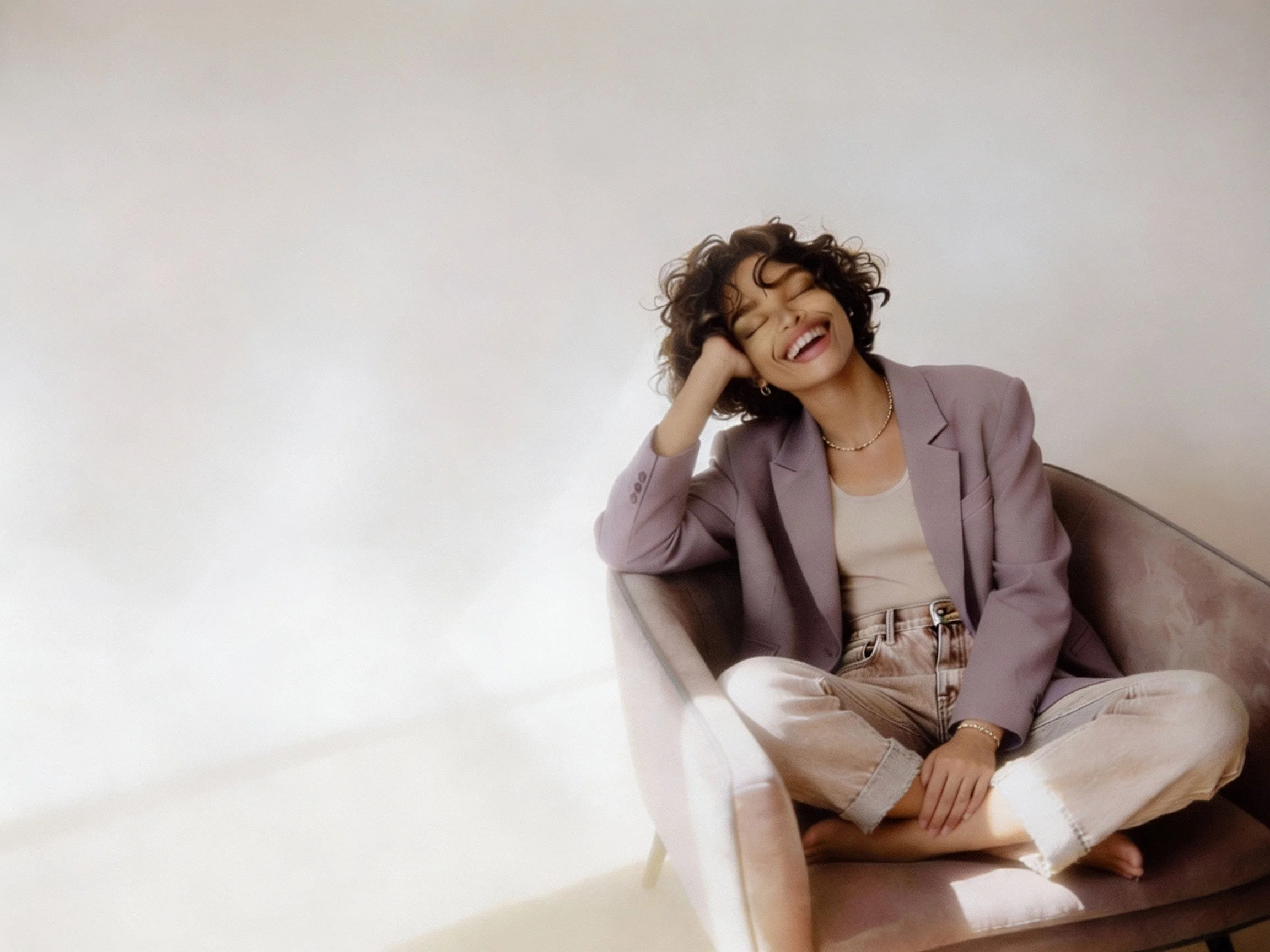 A woman with curly hair is seated against a plain white background, smiling and laughing with her eyes closed, wearing a light lavender blazer, beige top, and beige pants.