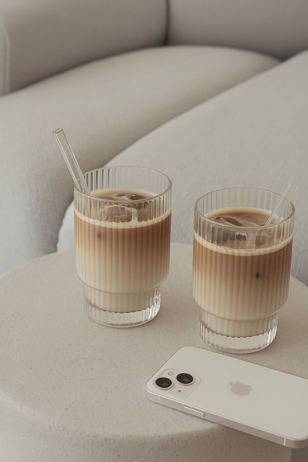 Two glasses of iced coffee with straws on a white table, next to a white iPhone, with a light-colored upholstered headboard in the background.
