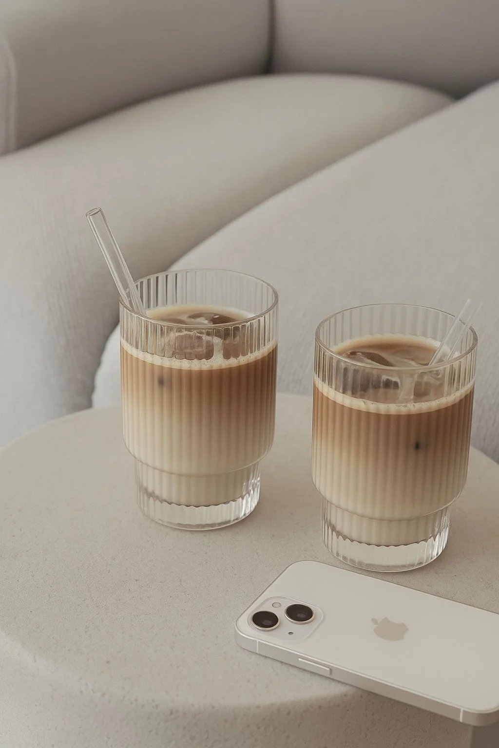 Two glasses of iced coffee with a pair of white headphones resting on their side on a beige surface.
