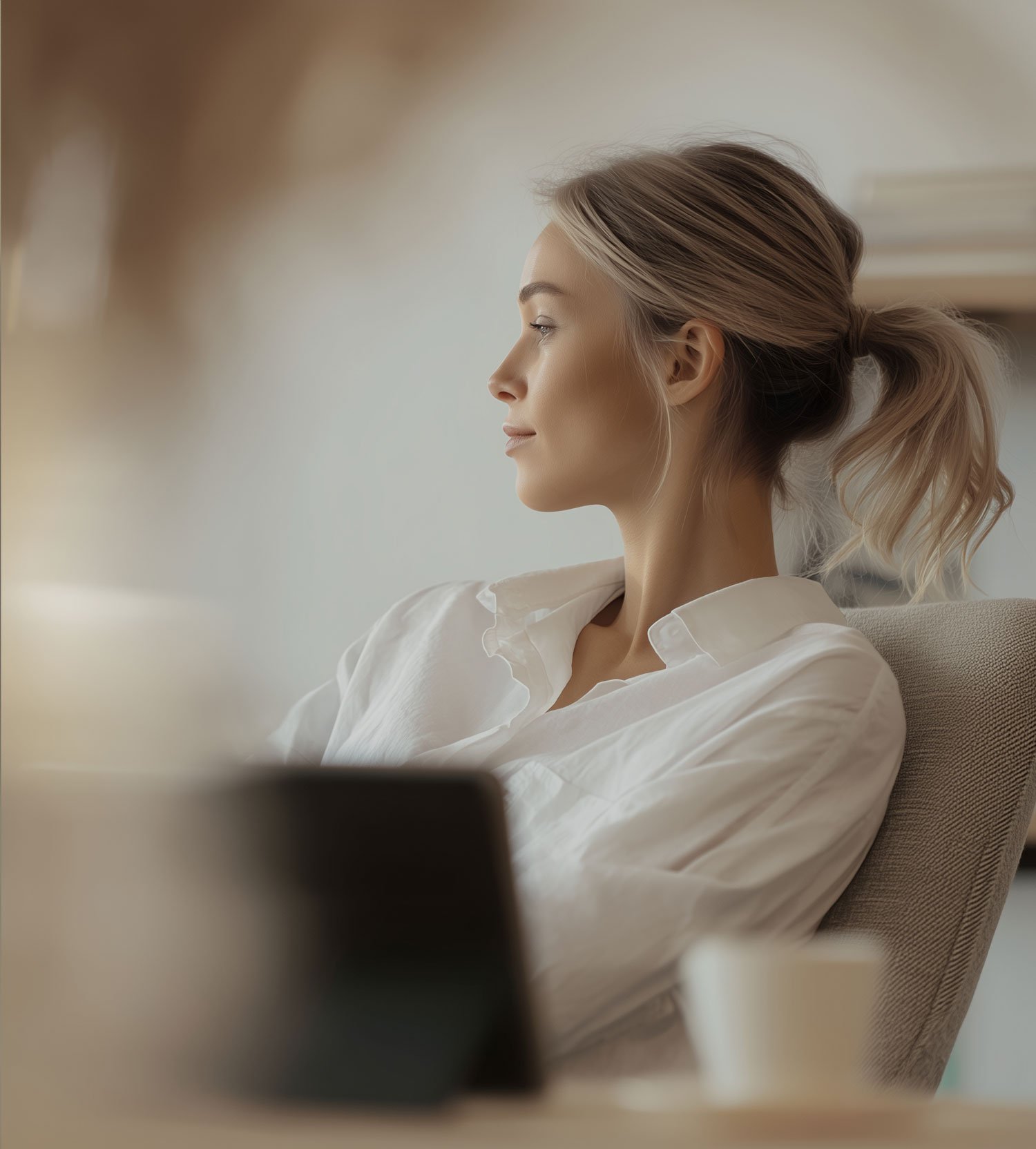 A woman in a white shirt sitting on a beige chair, looking to the left, with her hair tied back in a loose ponytail, in a softly lit room.