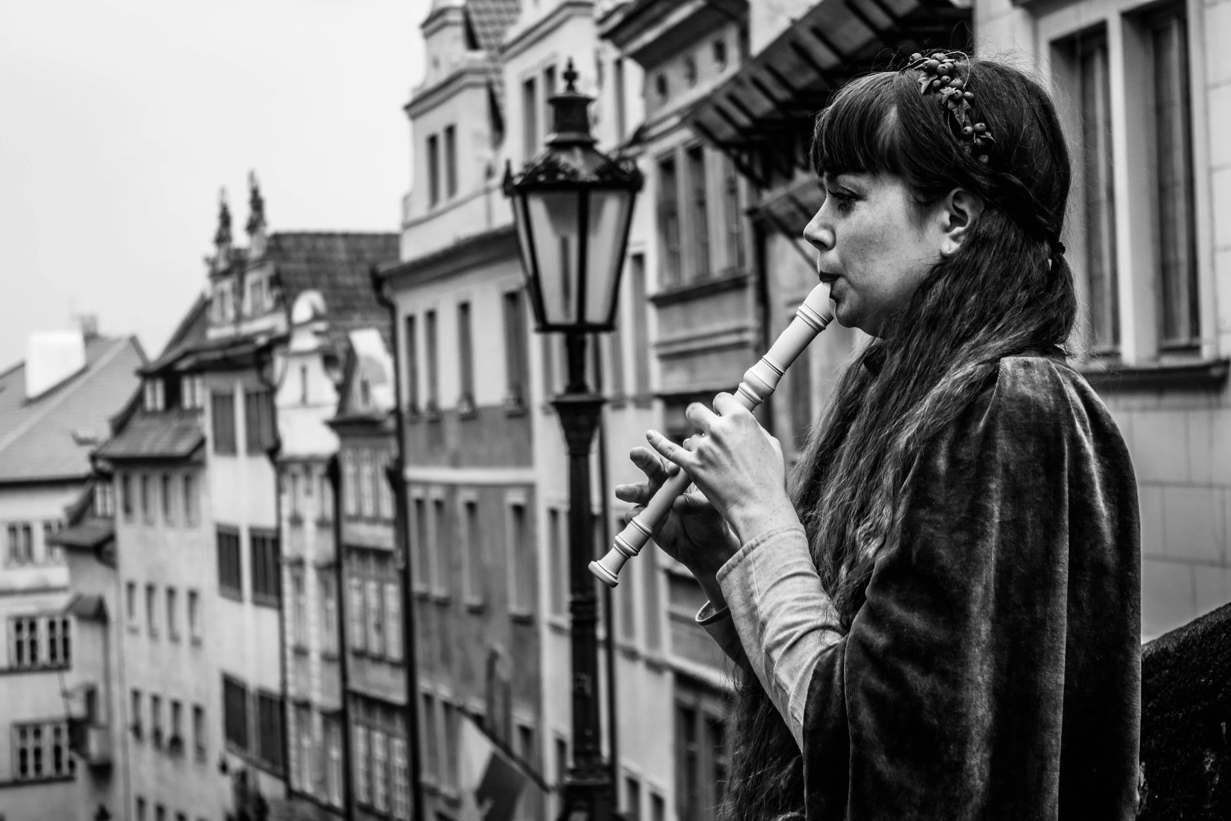 A woman playing a flute outdoors on a balcony with European-style buildings in the background, black and white photograph.