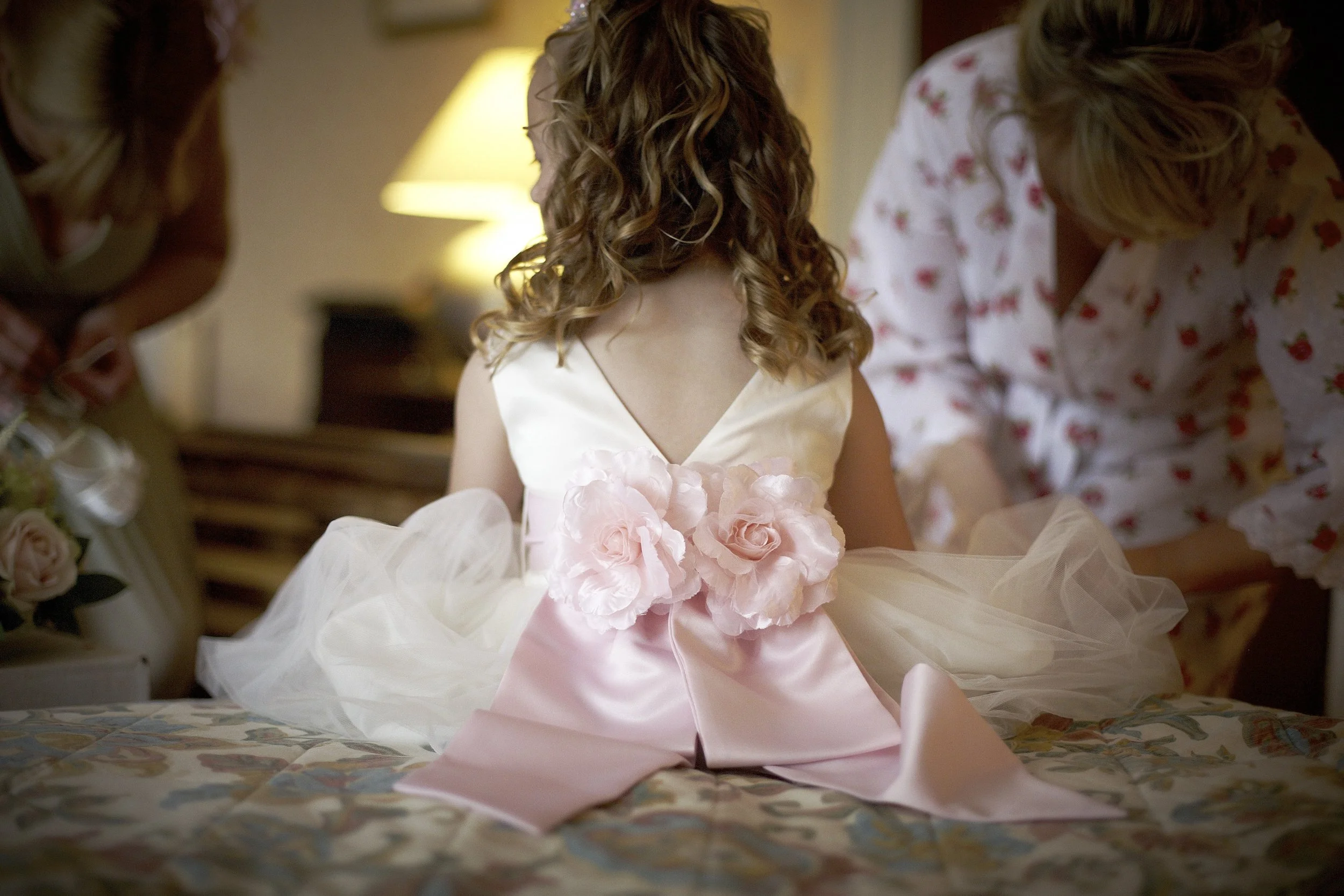 A young girl in a fancy pink dress with rose embellishments, sitting on a bed, while two women adjust or help her with her dress in a warmly lit bedroom.