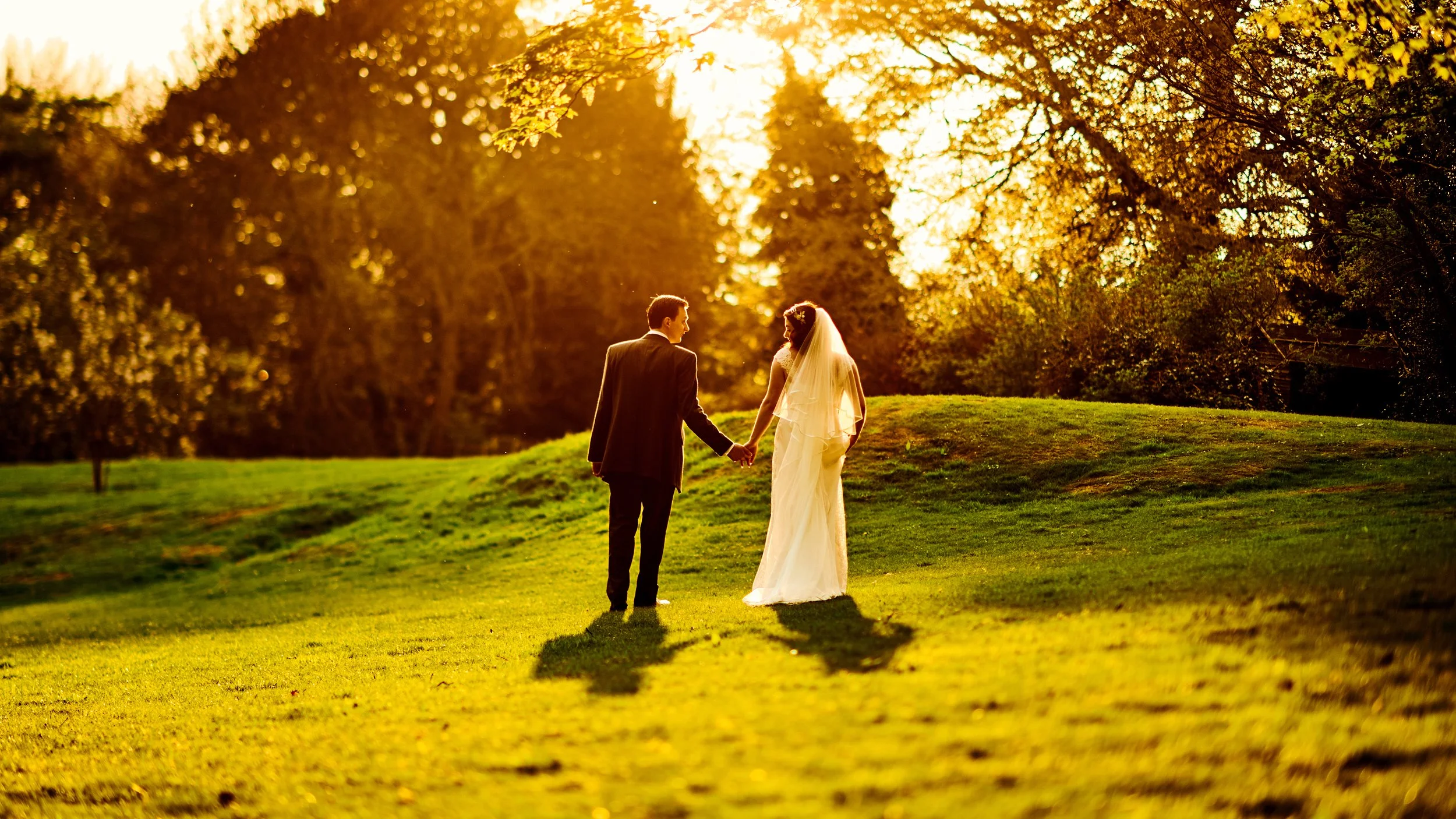 A bride and groom holding hands outdoors during sunset, walking across a grassy area with trees in the background.