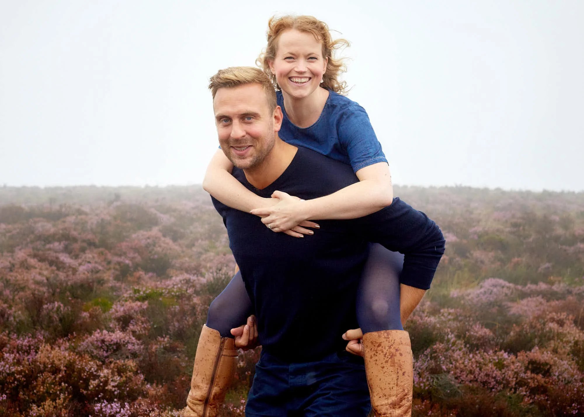 A woman giving a piggyback ride on a man in an outdoor field with purple flowers.