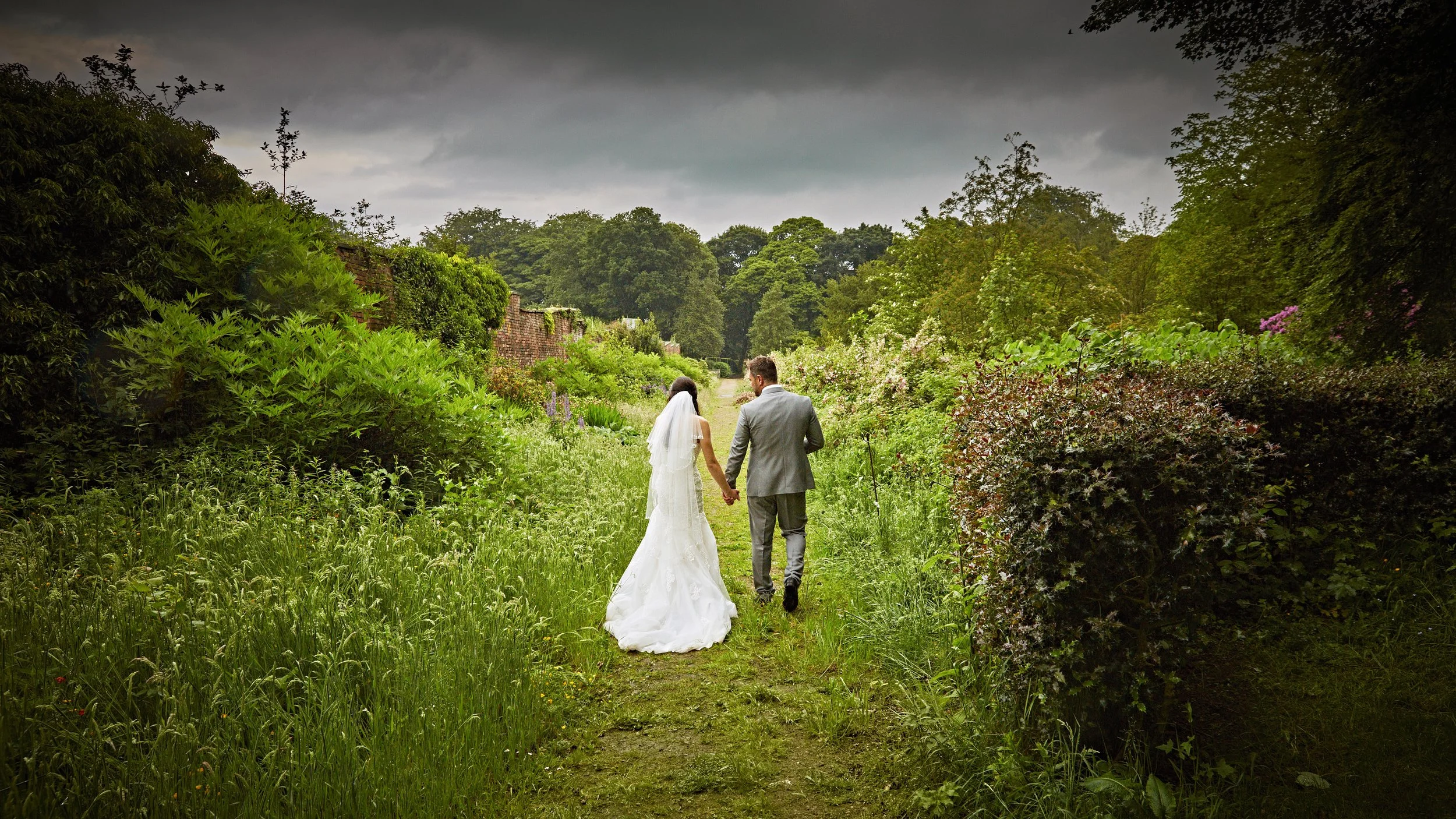 A bride and groom walking hand in hand through a lush green garden path on a cloudy day.