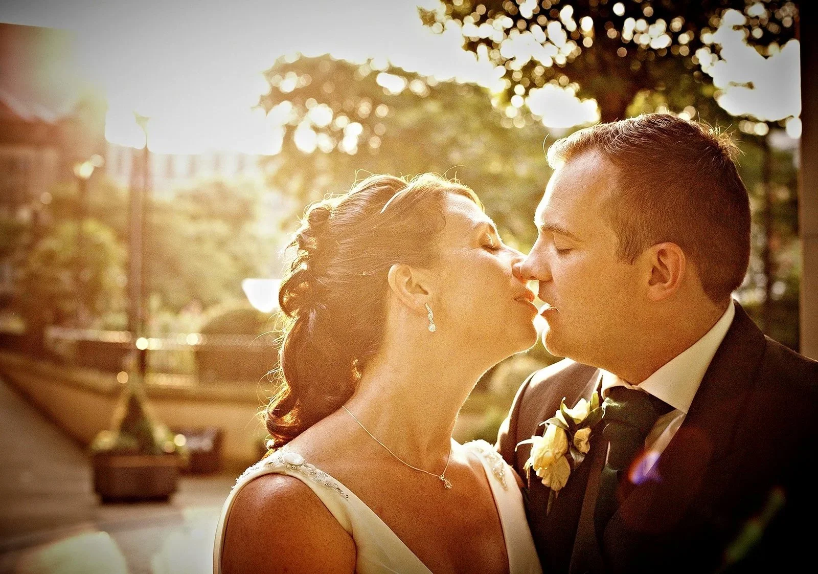 A bride and groom sharing a kiss during their wedding, with sunlight filtering through trees in the background.