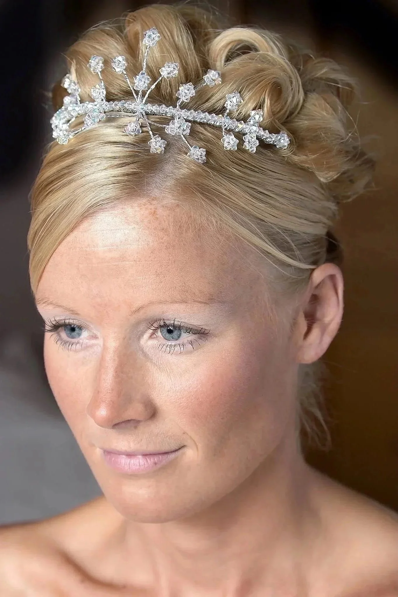 Close-up of a woman with blonde hair styled with a silver and crystal headpiece, smiling softly, with clear blue eyes and light makeup.