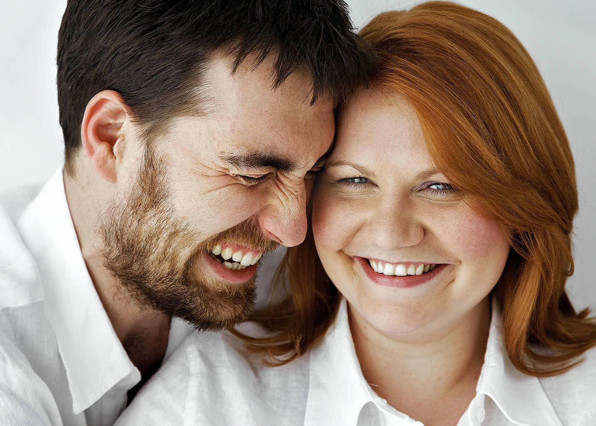 A close-up of a smiling man and woman with heads pressed together, both wearing white shirts, sharing a joyful moment.