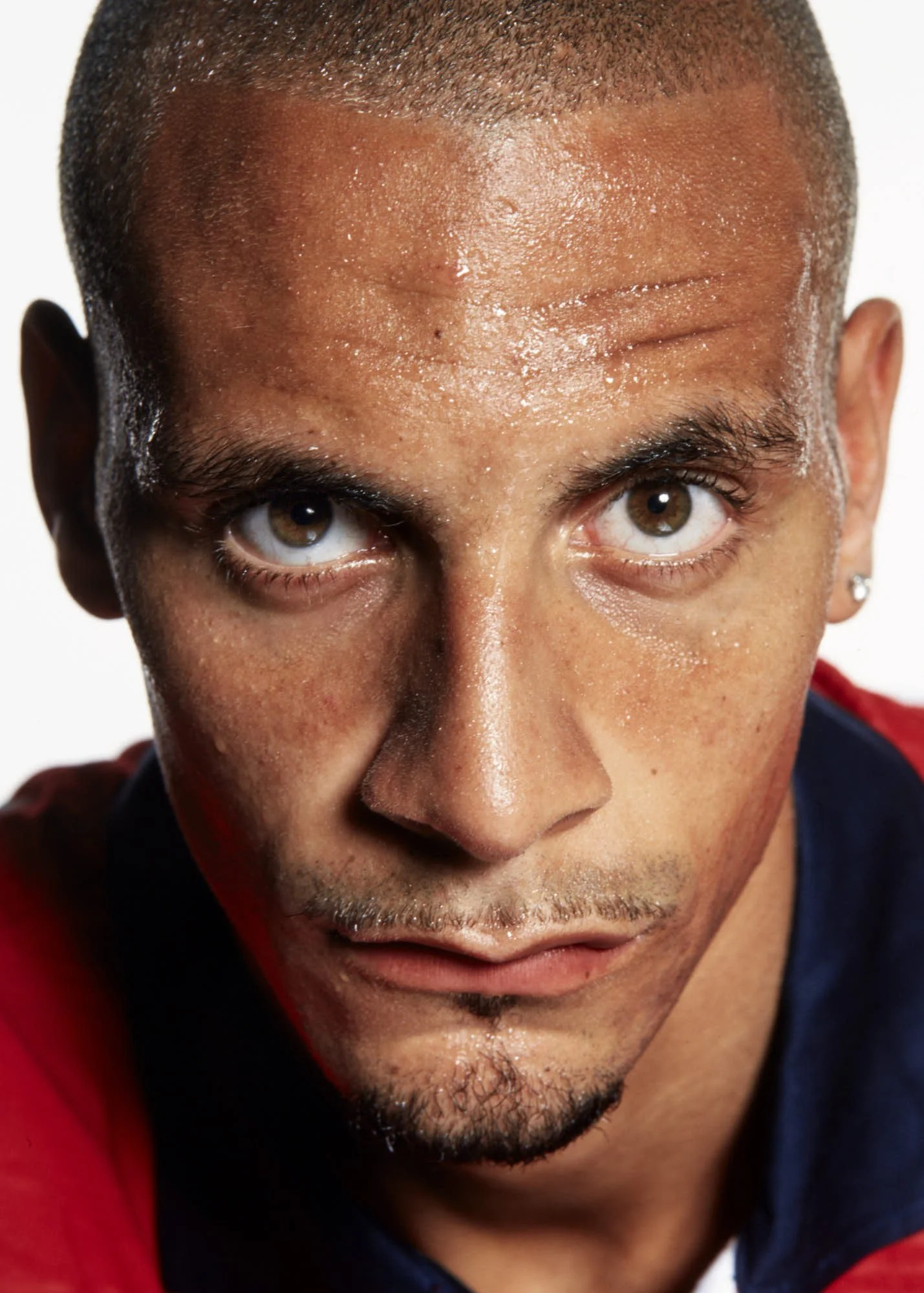 Close-up of a person's face with short hair, brown eyes, slightly furrowed brows, and a neutral expression, wearing a red and dark-colored hoodie.