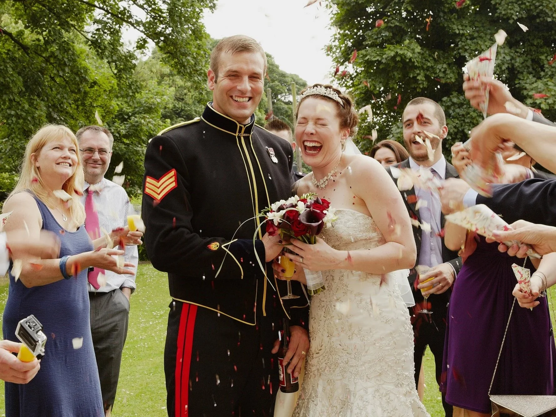 A wedding celebration outdoors with the bride and groom in the center, surrounded by cheering guests. The bride is holding a bouquet of red and white flowers, and the groom is wearing a formal military uniform. Celebratory showering and confetti are visible.