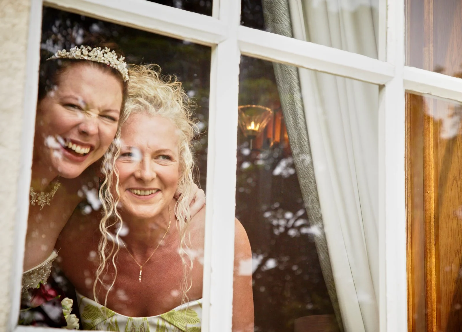 Two women smiling and looking through a window, one may be a bride with a floral headband, and the other an older woman, possibly a guest or family member, with curly blonde hair, in a cozy, warmly lit room.