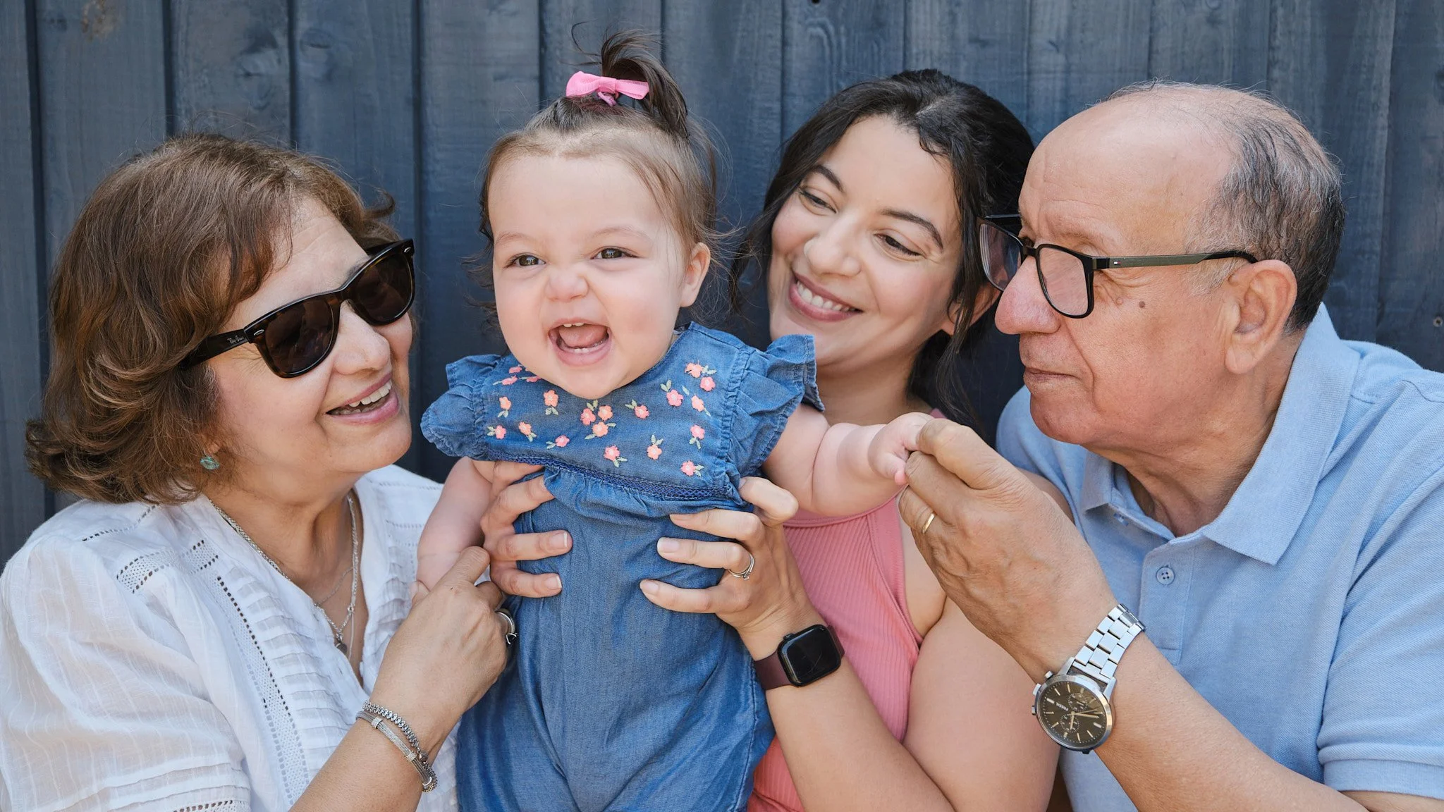 A group of five people, including two elderly adults, a young woman, a young girl, and a baby girl, smiling and enjoying each other's company outdoors against a dark wooden fence.