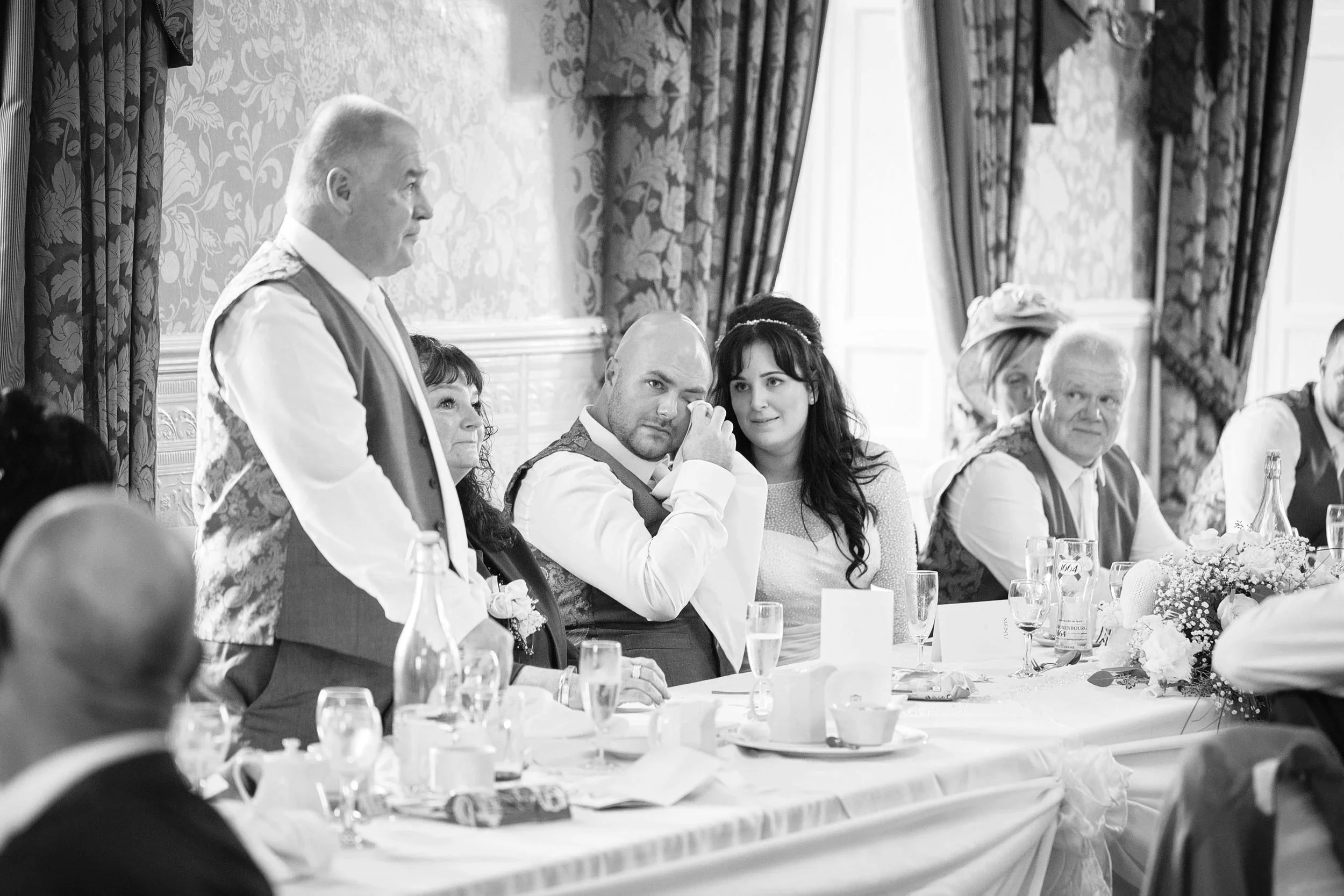 A black and white photo of a formal gathering with people seated at a long table, some wearing vests and white shirts, in a richly decorated room with floral patterned curtains and ornate wallpaper.