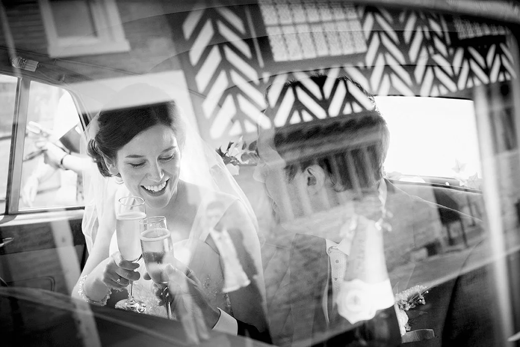 A bride and groom sitting inside a car, smiling, holding champagne glasses, seen through the car's window with reflections.
