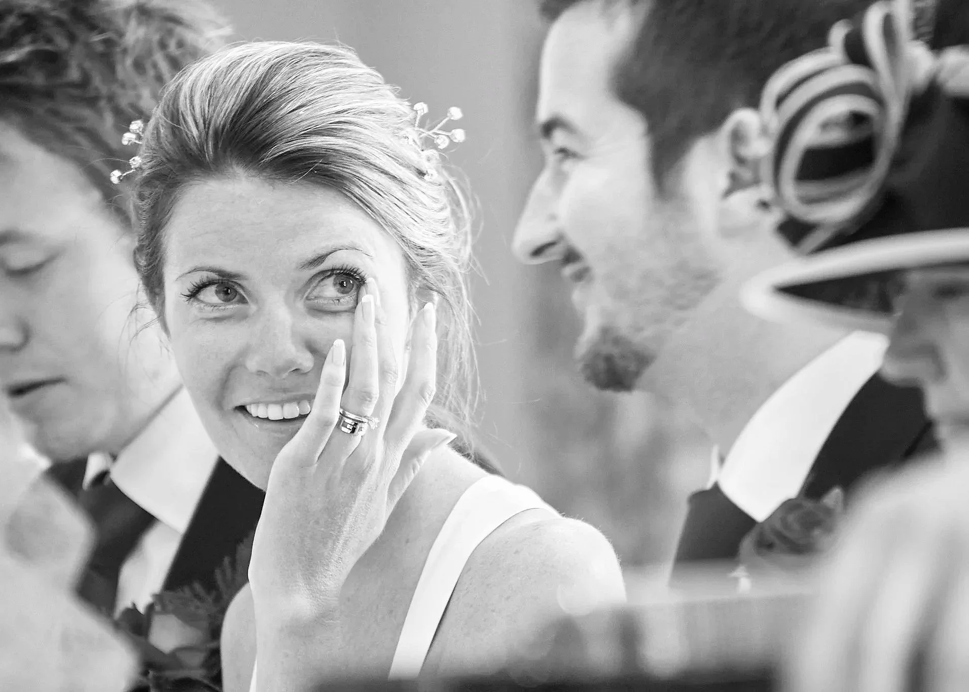 A wedding celebration with a bride in a wedding dress, surrounded by guests, whispering to her and smiling, in a candid black-and-white photo.