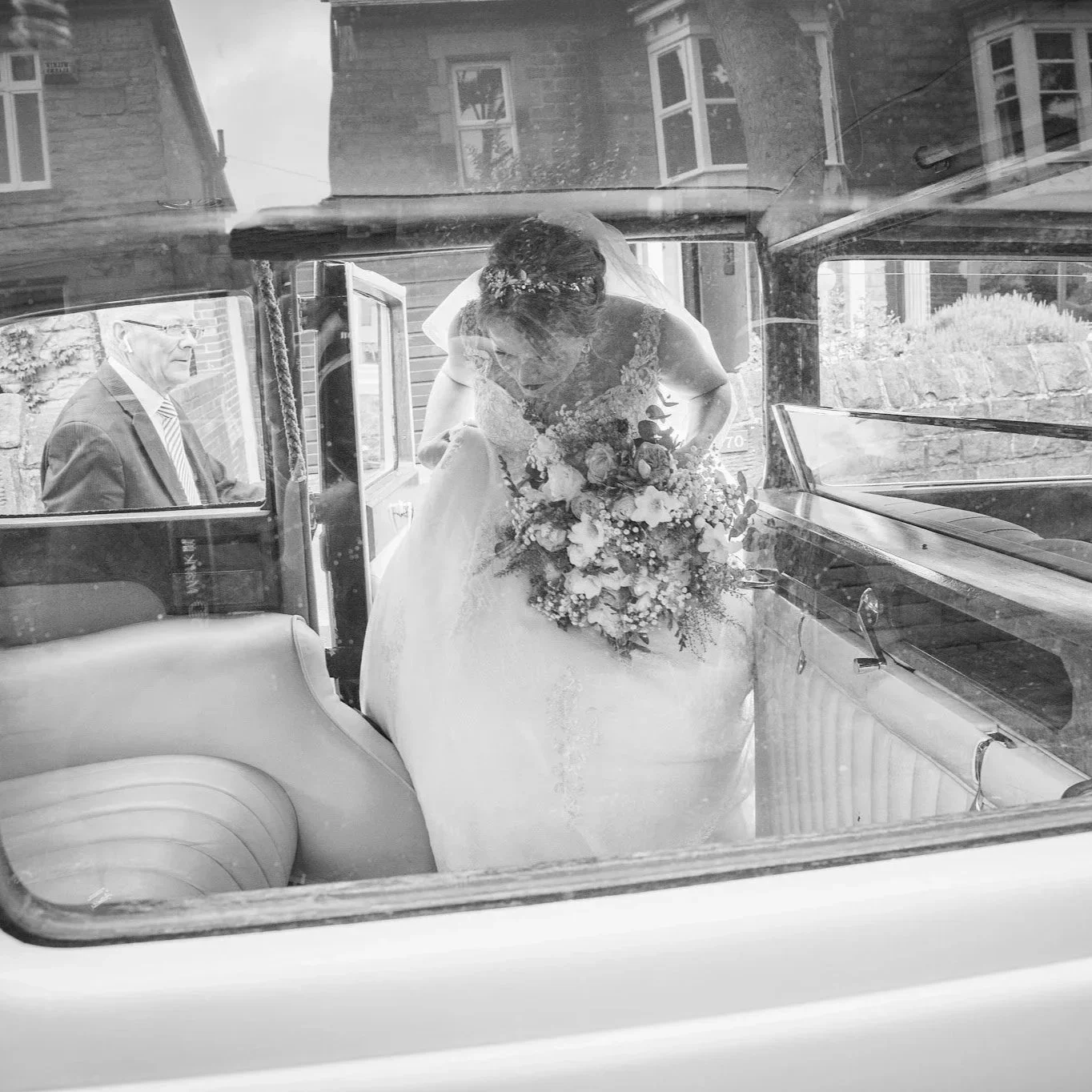 A bride in a wedding dress holding a large bouquet inside a vintage car, with an older man in a suit sitting nearby.
