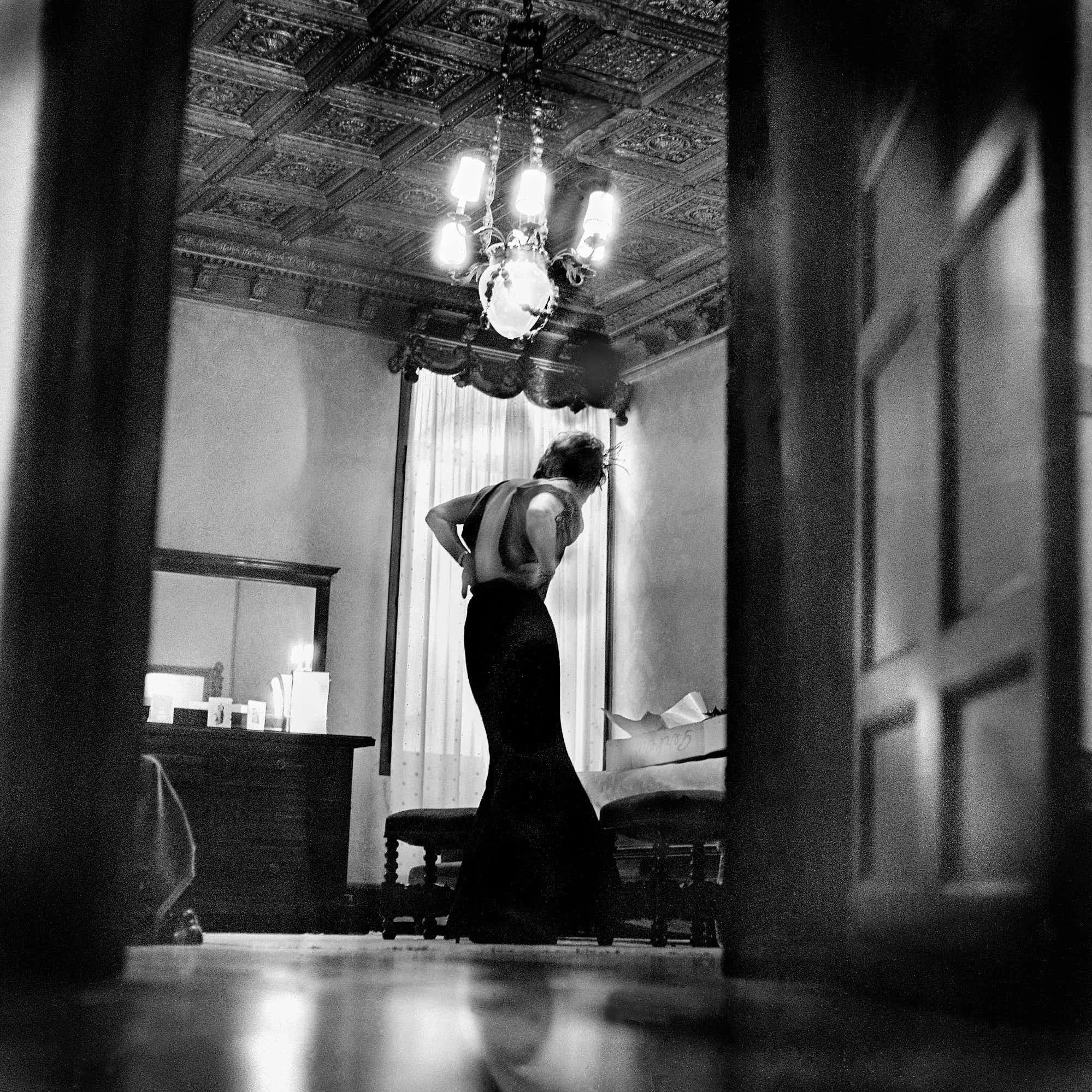 A person with curly hair leaning over a table with papers or artwork, in a room with ornate ceiling and chandelier, seen through a doorway with framed wall art.