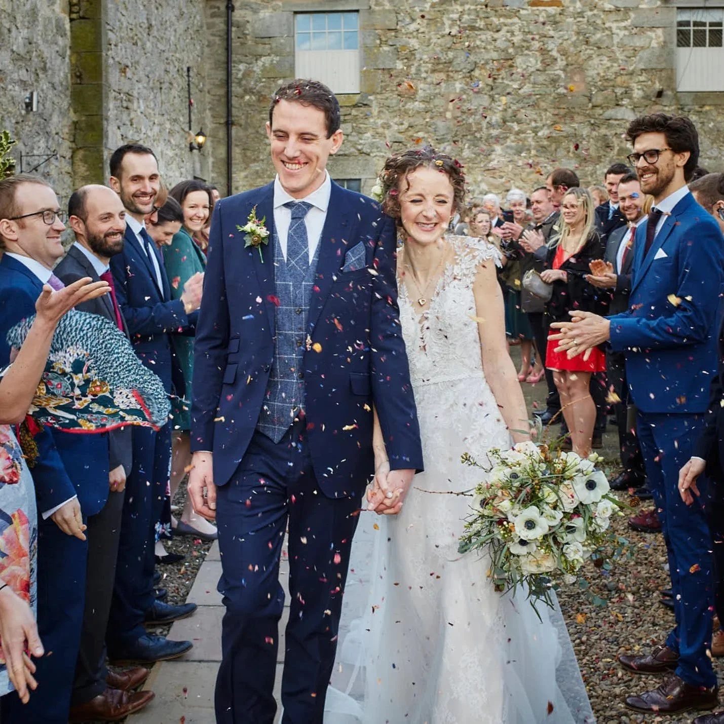 A newly married couple walking hand in hand, surrounded by friends and family, celebrating with flower petals and confetti outside a stone building.